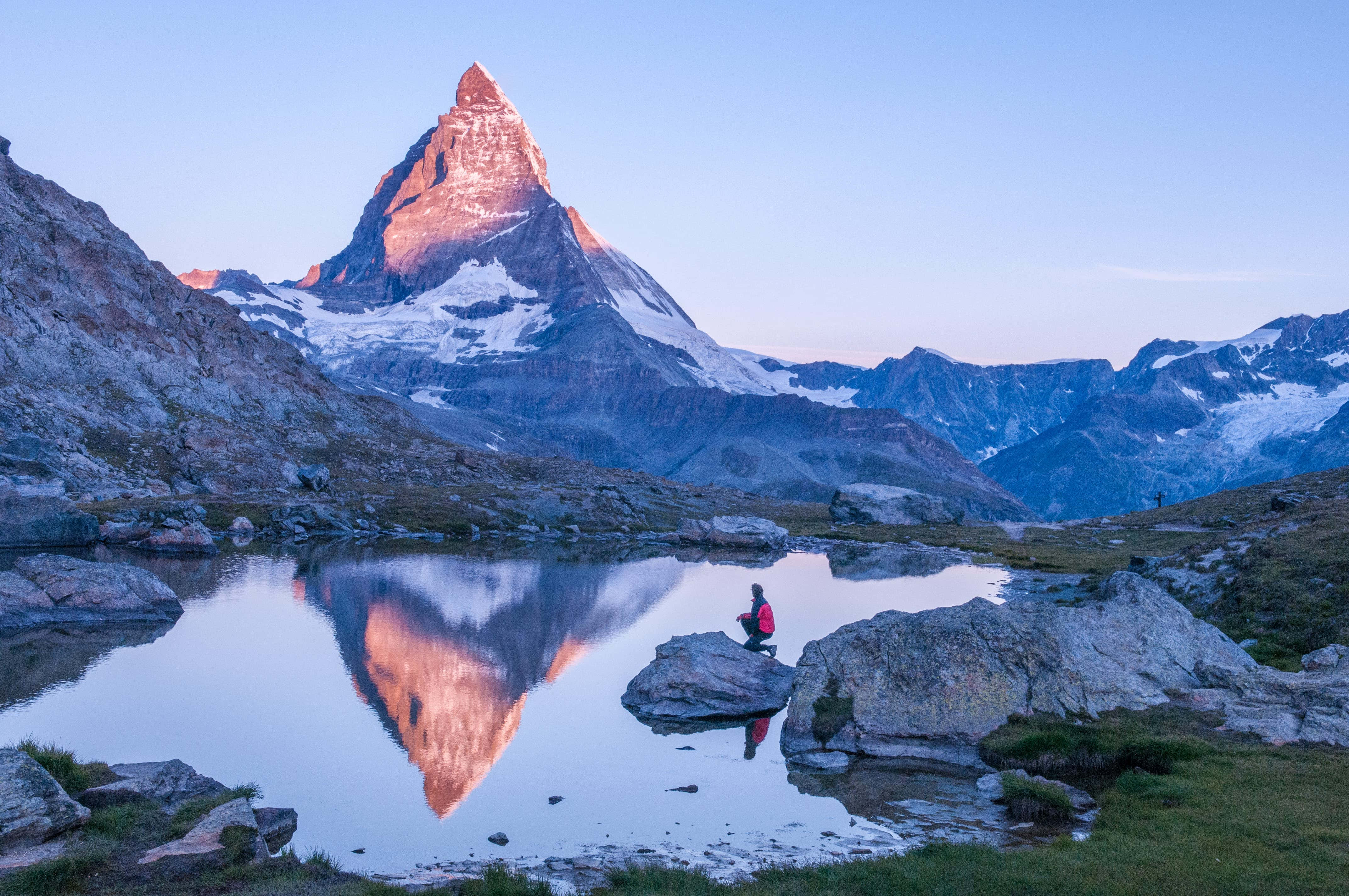 Switzerland 桌布 壁紙 — 一位孤獨的遠足者坐在寧靜高山湖邊的岩石上，欣賞馬特洪峰在日出時閃耀著粉紅色和金色的光芒。
