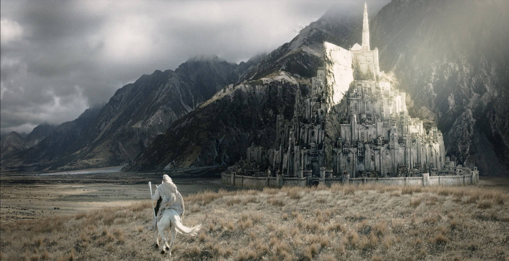 Lord Of The Rings Landscape 桌布 壁紙 — 一位孤獨的騎手騎著白馬，在暴風雨的天空下，奔馳過荒蕪的平原，朝向一座雄偉的石頭堡壘，該堡壘坐落於山頂之上。