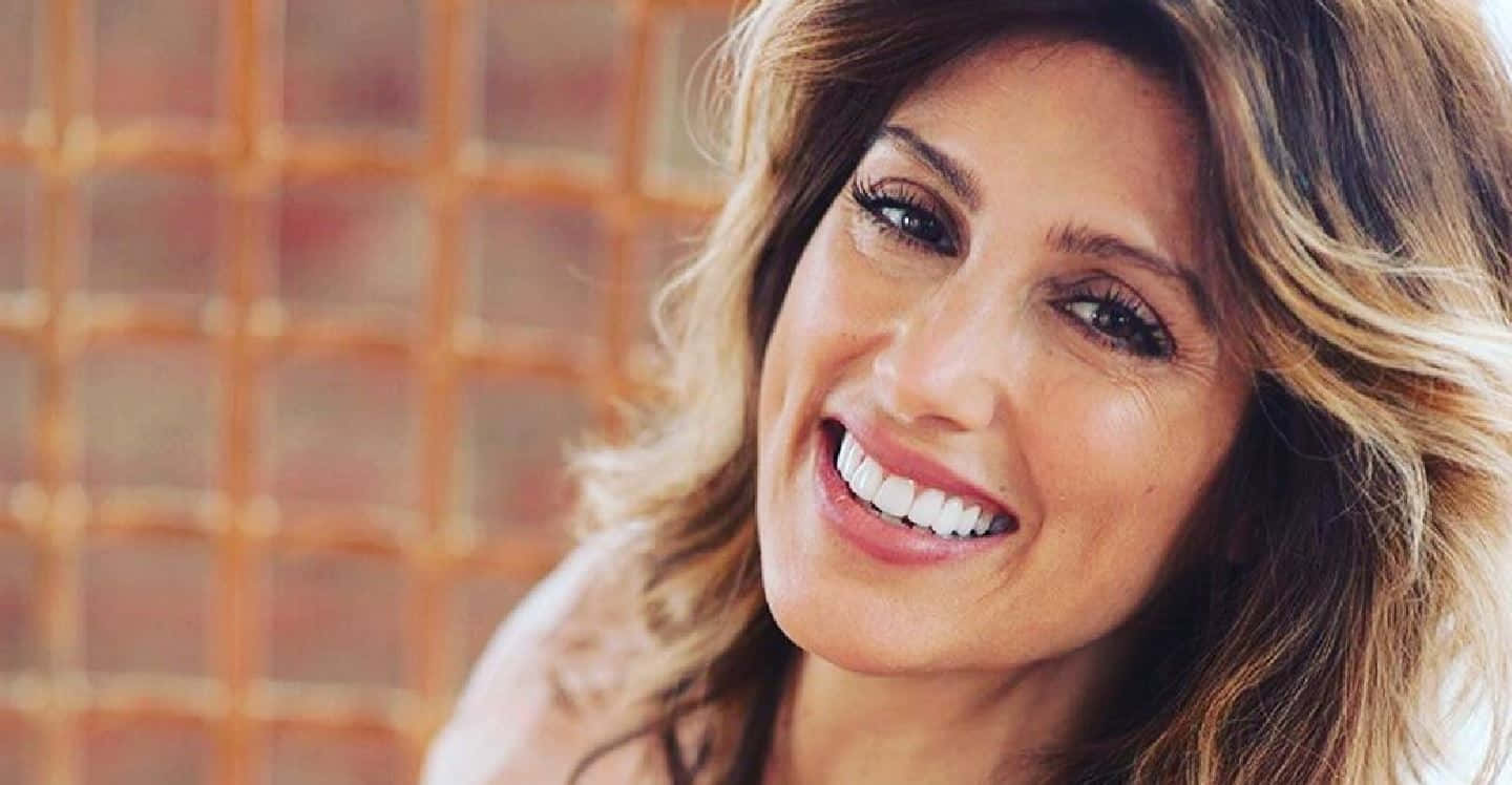 Jennifer Esposito 桌布 壁紙 — 一位容光煥發、擁有一頭波浪金髮的女性，在暖色調的瓷磚背景前燦爛微笑。