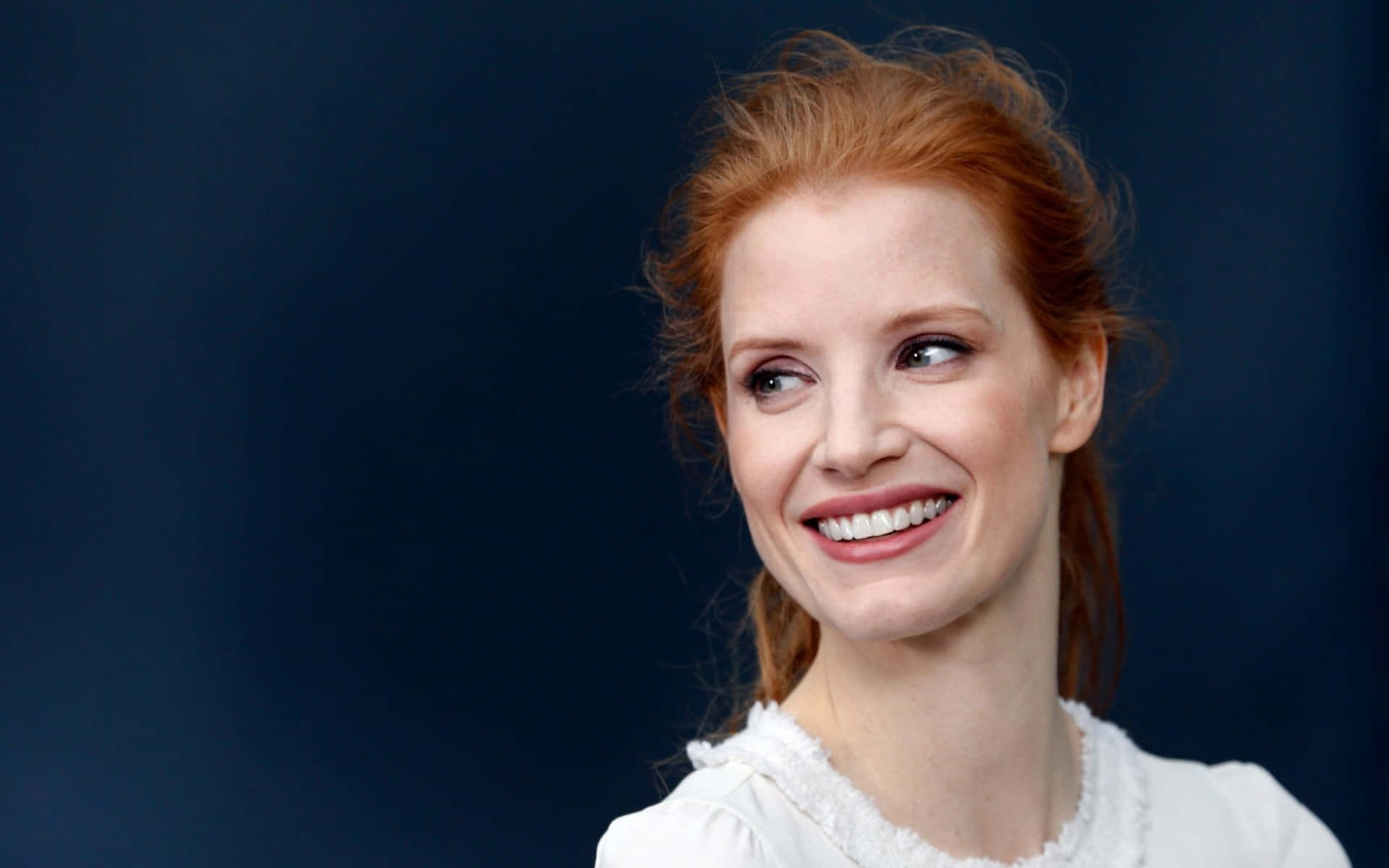 jessica chastain 桌布 壁紙 — 一位容光煥發、擁有火紅頭髮的女性在深藍色攝影棚背景下溫暖地微笑。