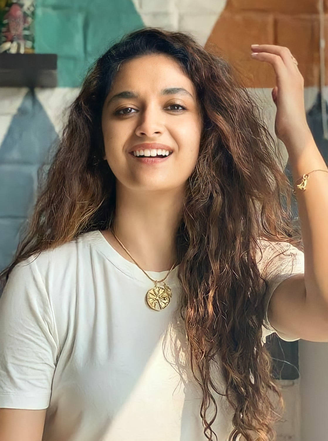 Tamil Actors HD 桌布 壁紙 — 一位容光煥發的女士，長著波浪形棕色長髮，在色彩繽紛的幾何牆壁前微笑著觸摸自己的頭髮。