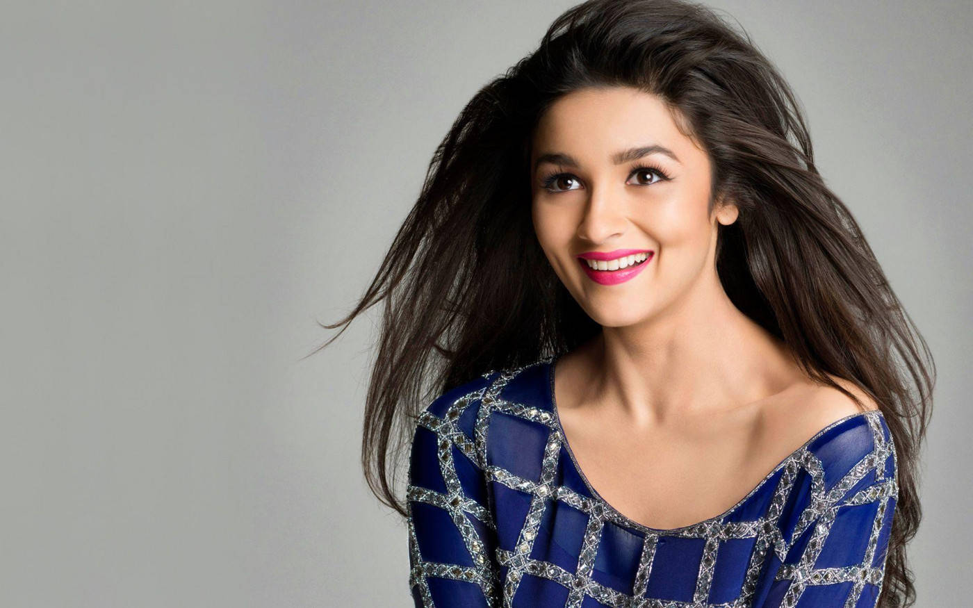Alia Bhatt HD 桌布 壁紙 — 一位容光煥發的女性，擁有飄逸的深色頭髮和亮粉紅色唇膏，穿著藍色圖案上衣，在灰色中性背景前溫暖地微笑。