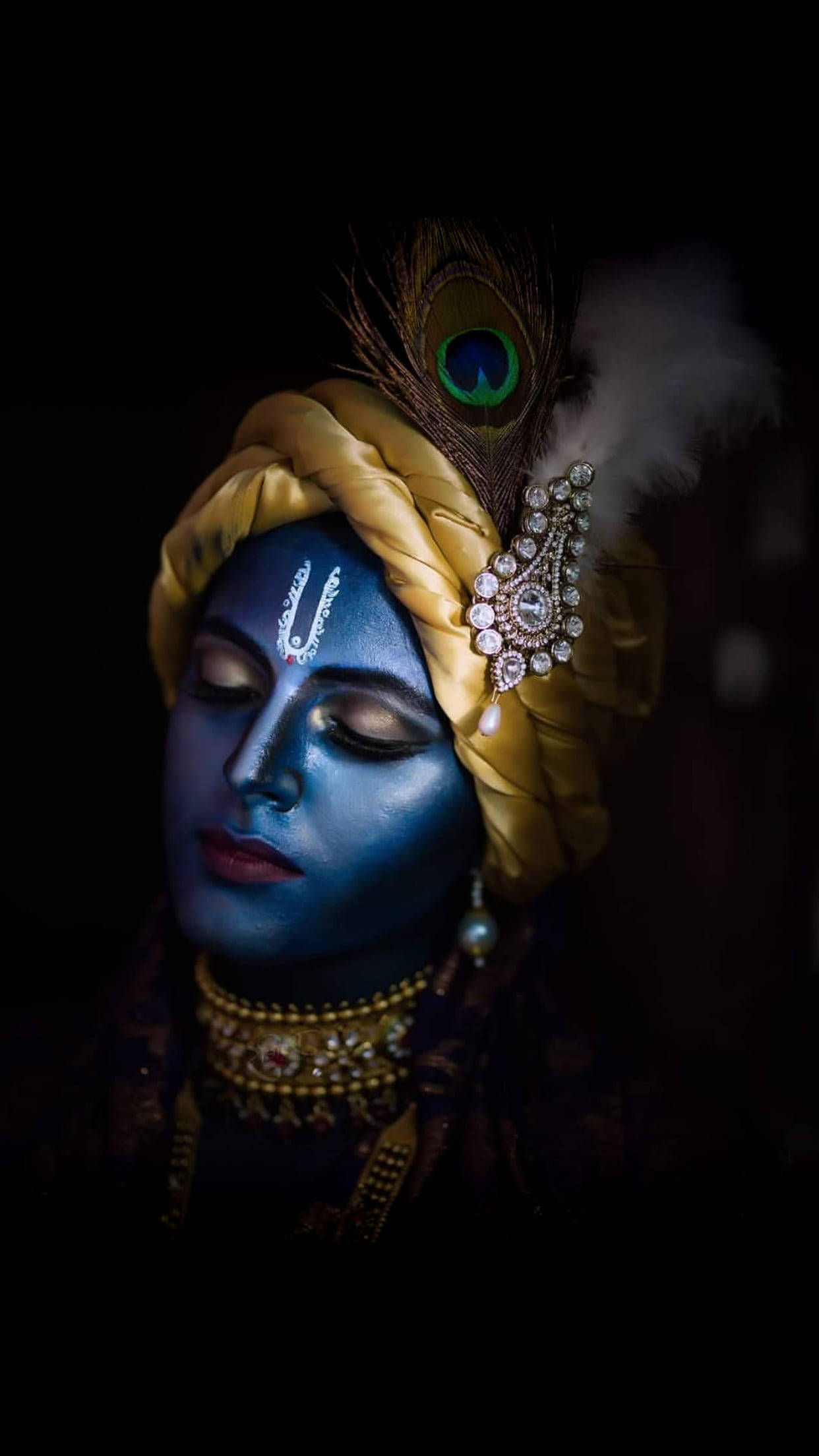 krishna bhagwan 桌布 壁紙 — 一位寧靜的藍皮膚神祇，閉著雙眼，佩戴金色珠寶和孔雀羽毛冠冕，背景為深色。