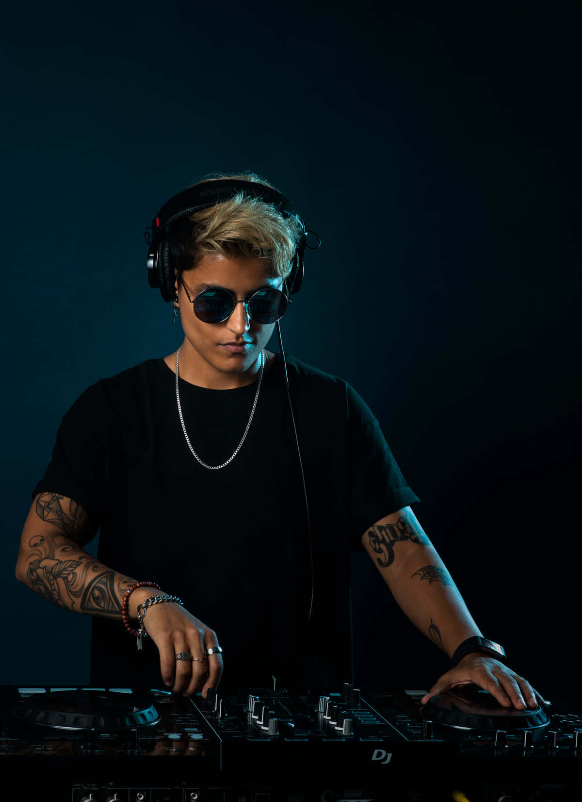 DJ 桌布 壁紙 — 一位專注的DJ，帶有紋身和太陽眼鏡，在戲劇性的藍色燈光下，在黑暗背景前操作唱盤。
