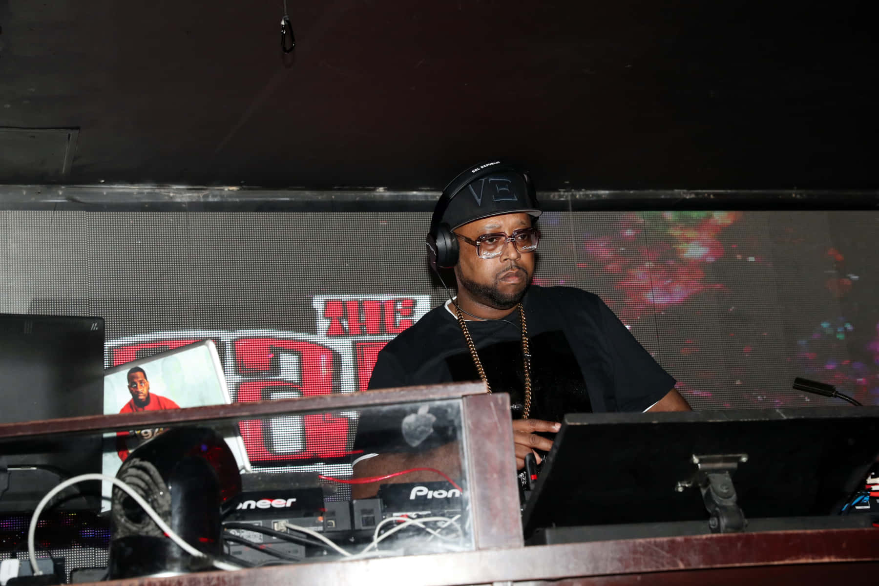 DJ 桌布 壁紙 — 一位專注的DJ戴著黑色鴨舌帽和眼鏡，在夜總會色彩繽紛的舞台燈光下操作唱盤。
