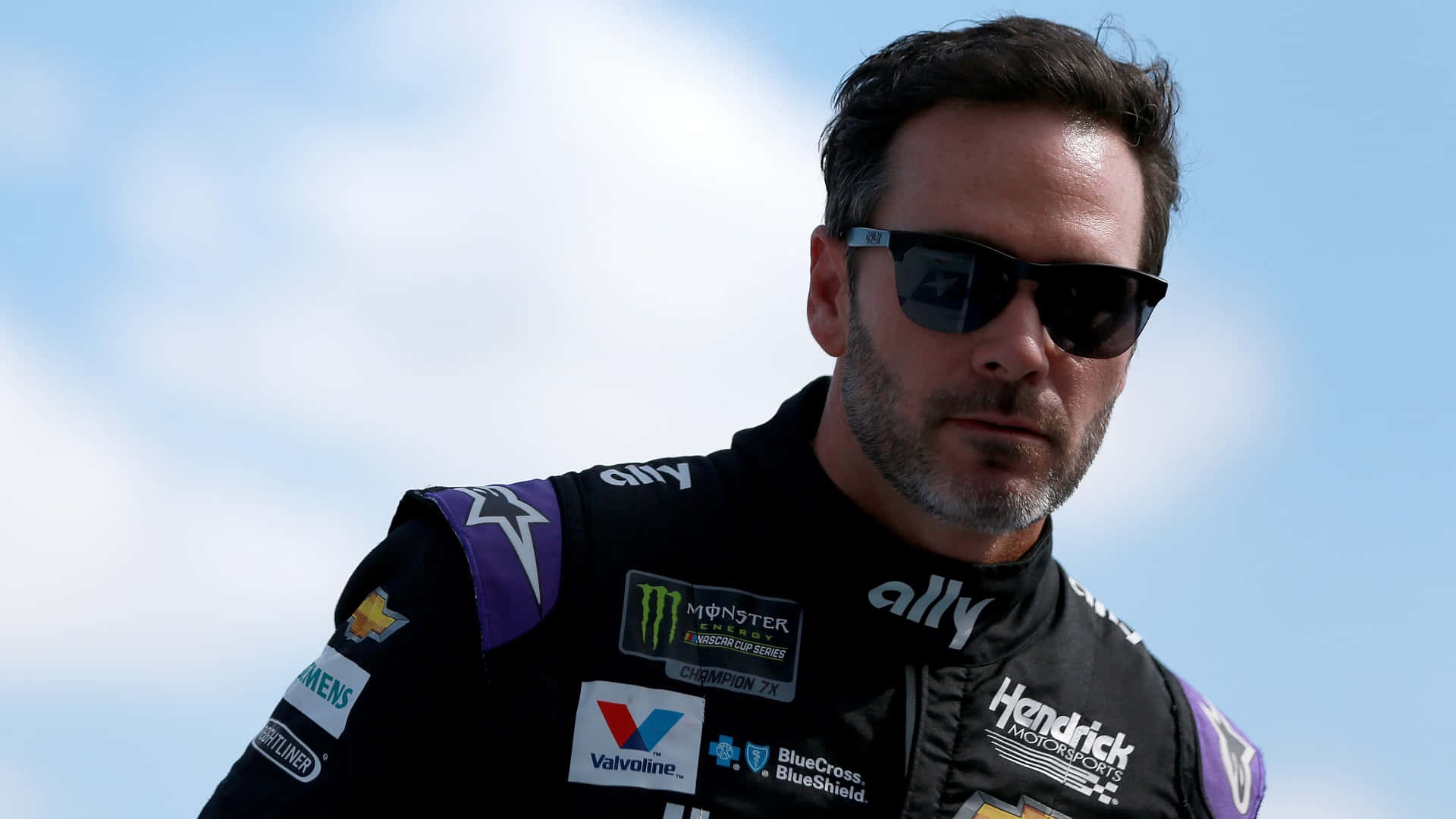 Jimmie Johnson 桌布 壁紙 — 一位專注的NASCAR車手穿著黑色和紫色的賽車服，帶有贊助商標誌，站在明亮的藍天和散落的雲朵前。