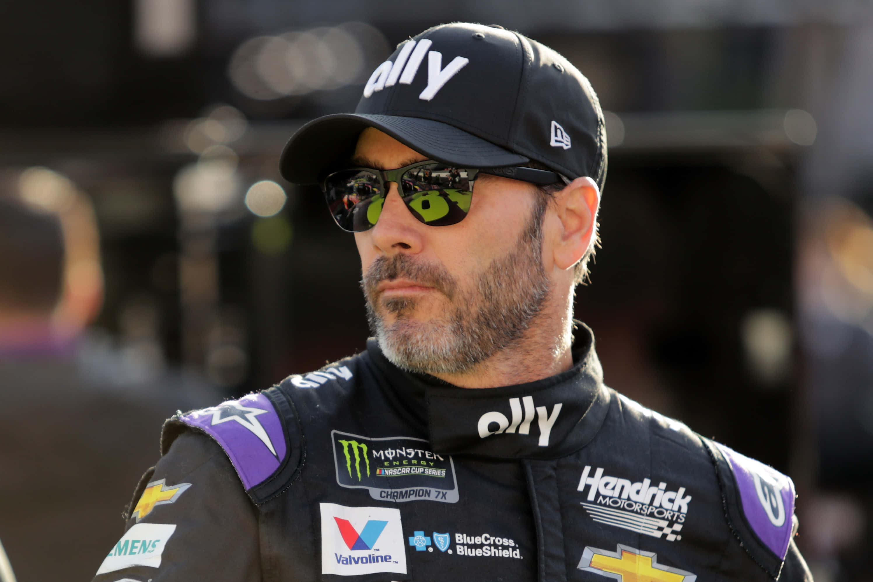 Jimmie Johnson 桌布 壁紙 — 一位專注的NASCAR車手穿著黑色賽車服和帽子，戴著綠色太陽眼鏡，在日間活動中凝視著。