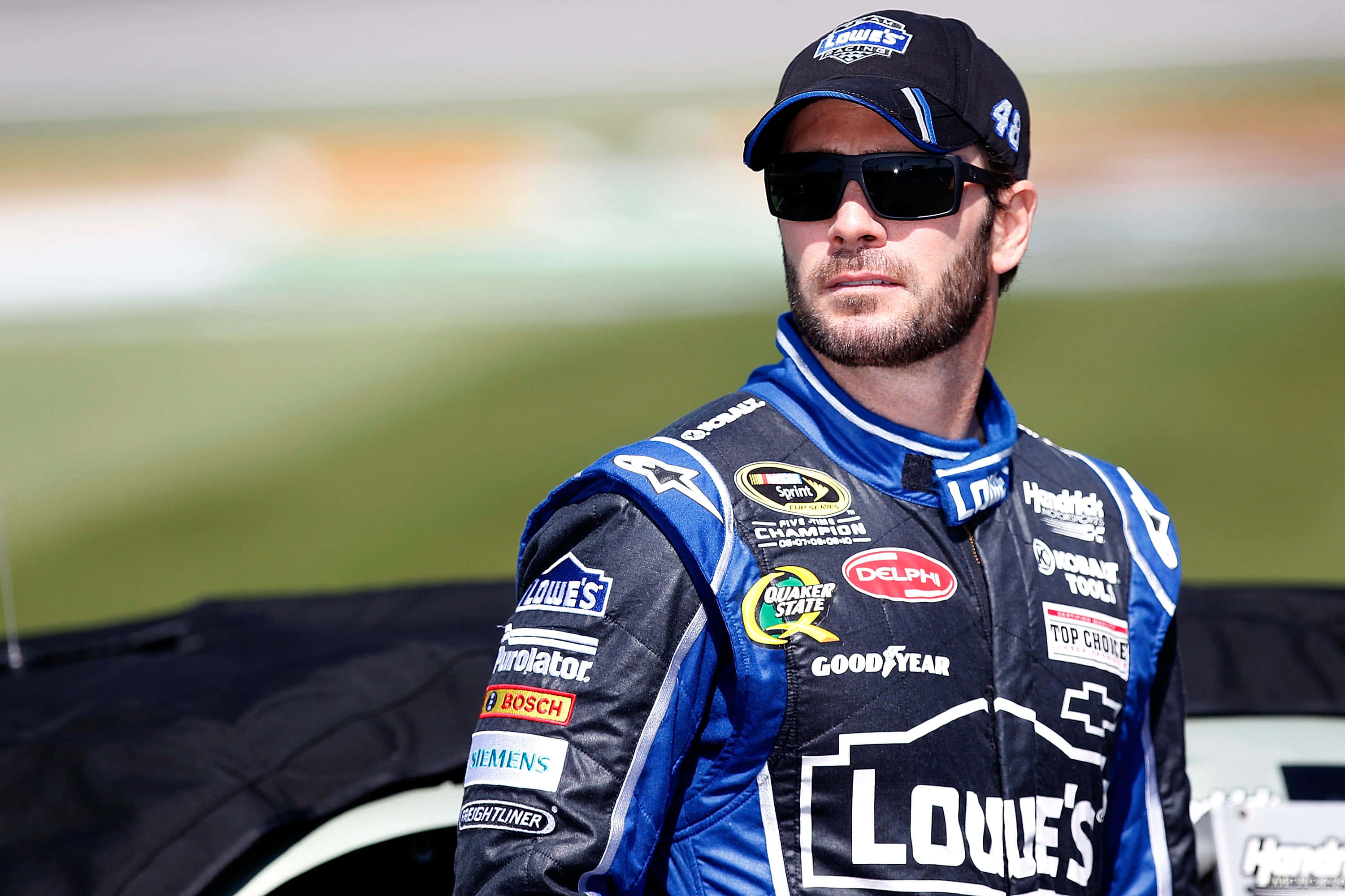 Jimmie Johnson 桌布 壁紙 — 一位專注的NASCAR車手身穿藍黑賽車服，在明亮的日光下站在他的賽車旁邊。