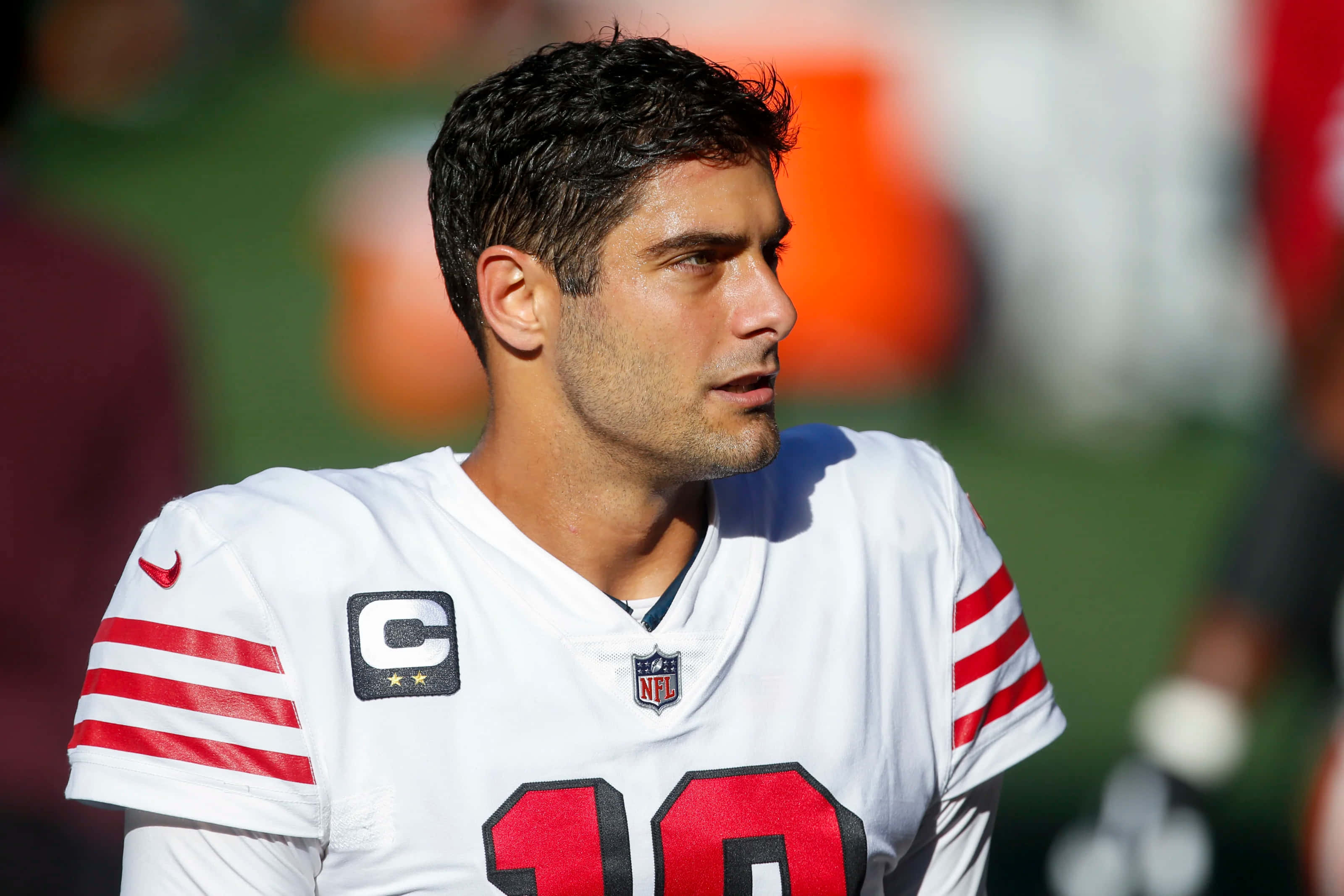 Jimmy Garoppolo 桌布 壁紙 — 一位專注的NFL四分衛，身穿帶有紅色條紋和10號的白色球衣，站在陽光普照的球場上。