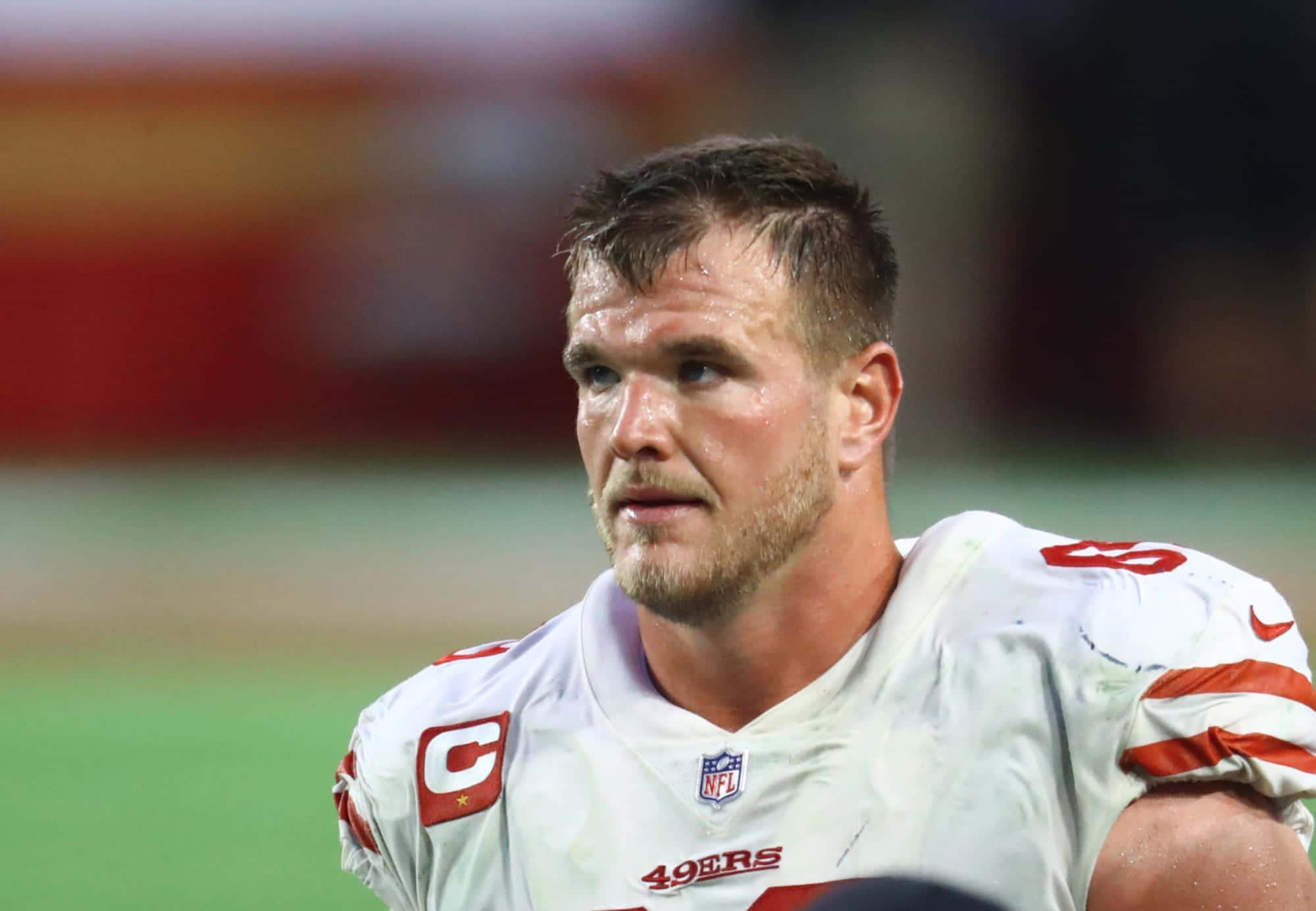 Mike Mcglinchey 桌布 壁紙 — 一位專注的NFL四分衛，身穿白色49ers球衣，帶有隊長'C'臂章，在夜間比賽中於球場燈光下專注地凝視。