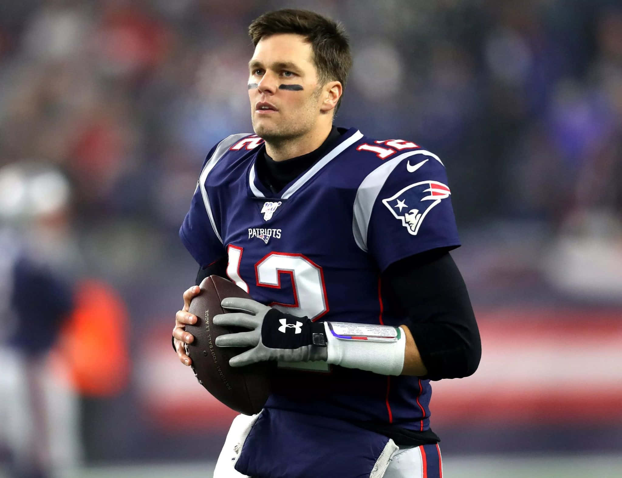 湯姆·布雷迪 桌布 壁紙 — 一位專注的NFL四分衛身穿新英格蘭愛國者隊制服，在球場燈光下持球。