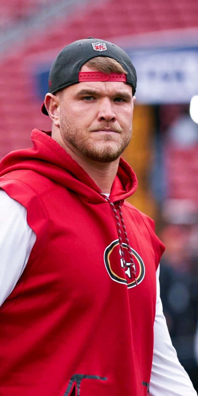 Mike Mcglinchey 桌布 壁紙 — 一位專注的NFL球員穿著紅色無袖連帽衫和黑色帽子，站在球場上，身後是模糊的紅色座位。