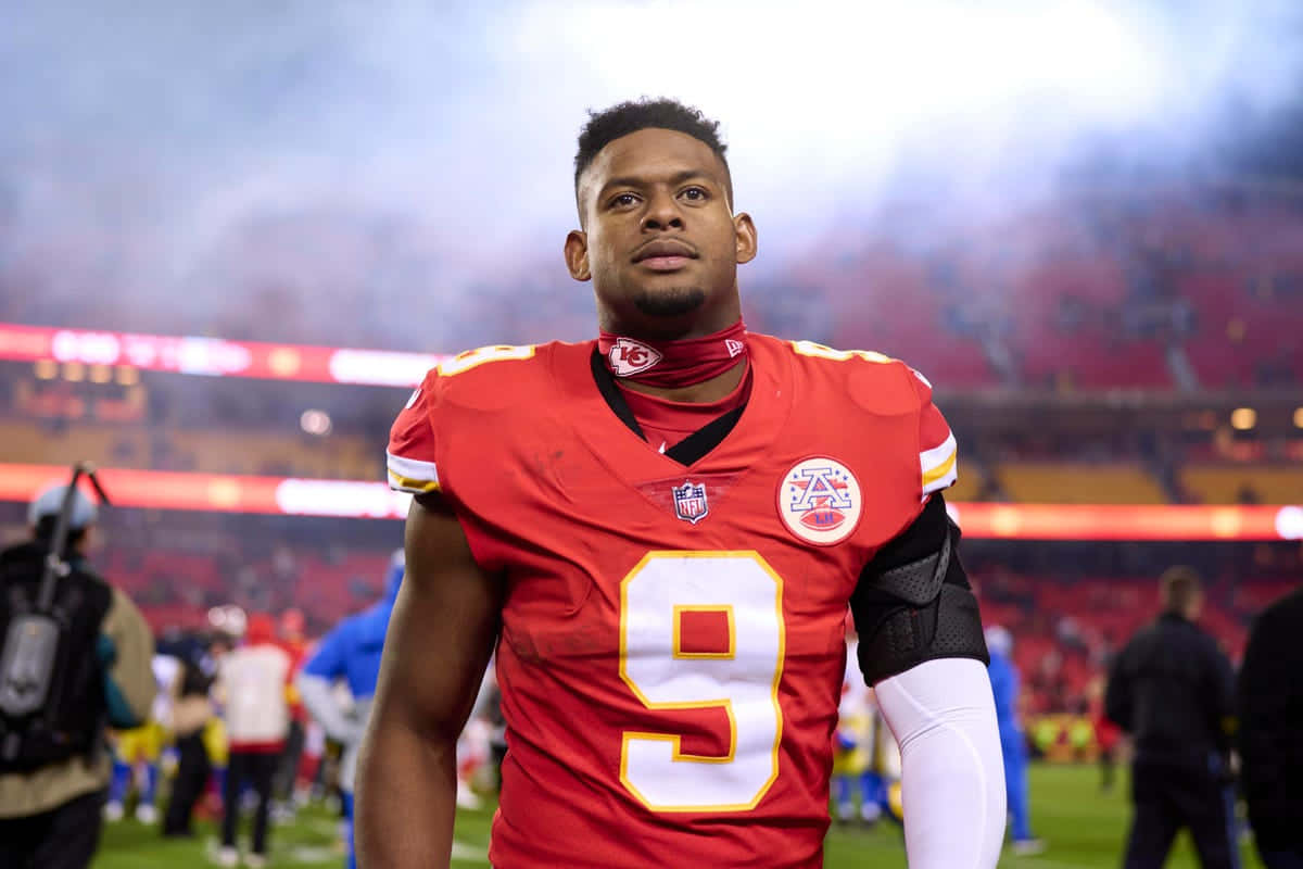 Juju Smith Schuster 桌布 壁紙 — 一位專注的NFL球員身穿紅色9號球衣，在明亮的燈光下站在煙霧繚繞的體育場上。
