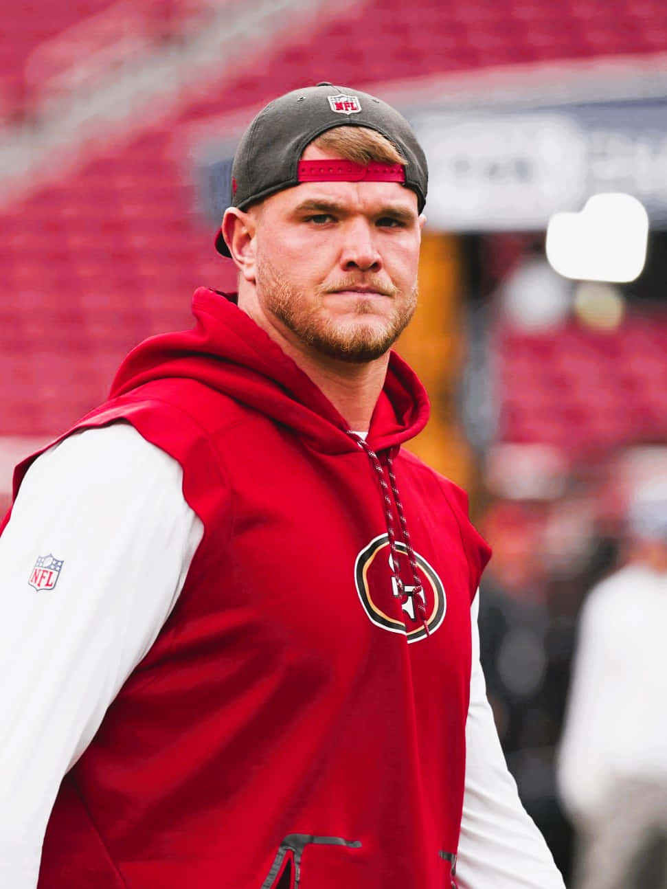Mike Mcglinchey 桌布 壁紙 — 一位專注的NFL球員身穿紅色三藩市49人連帽衫和帽子，站在球場上，背景是模糊的紅色座位。