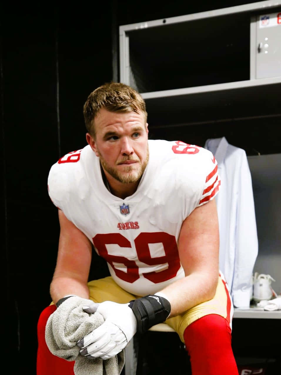 Mike Mcglinchey 桌布 壁紙 — 一位專注的三藩市49人美式足球員，身穿69號球衣，坐在燈光昏暗的更衣室裡，拿著毛巾。