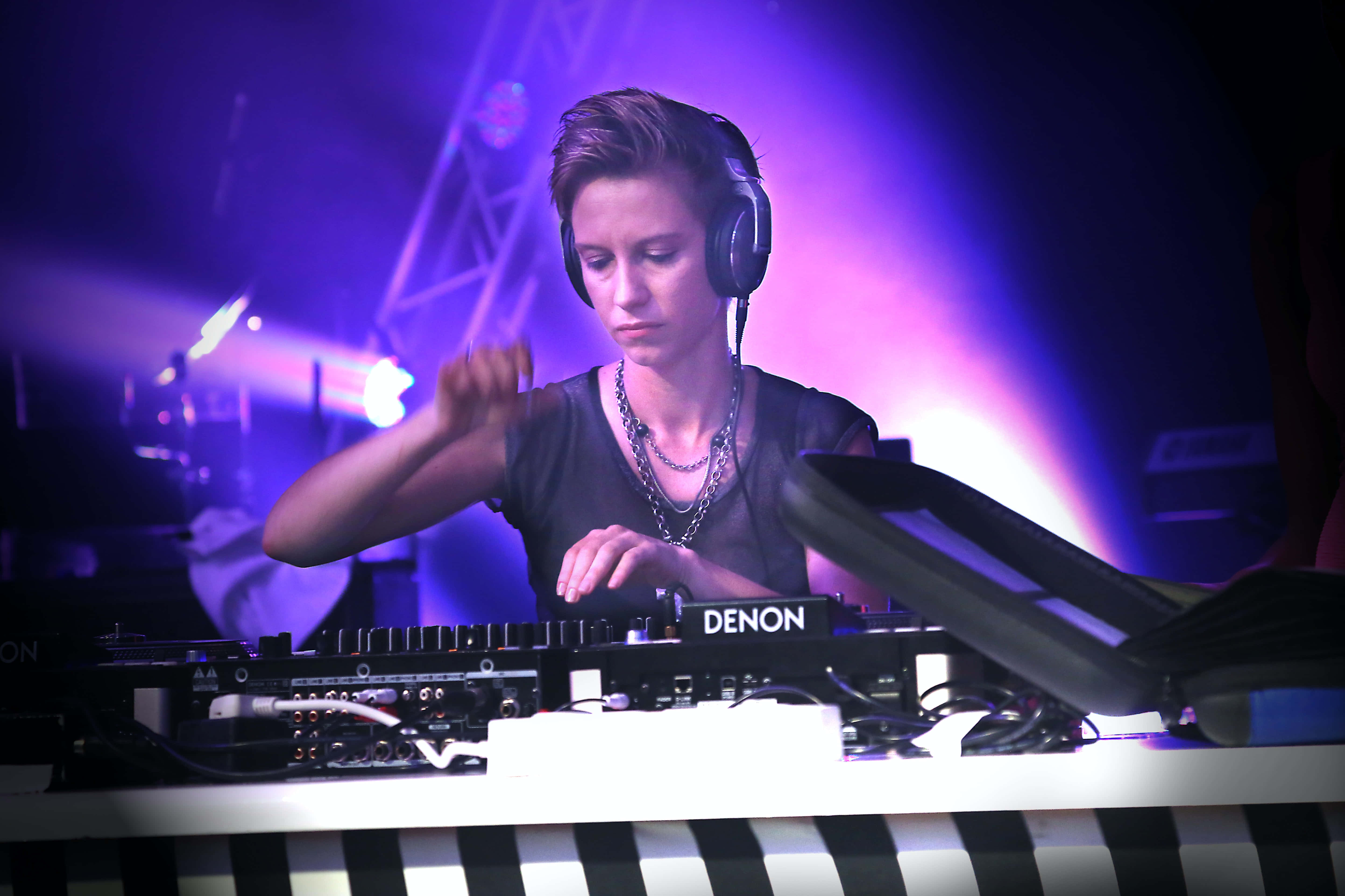 music festival 桌布 壁紙 — 一位專注的女DJ在充滿活力的紫色舞台燈光下操作Denon混音器的控制裝置。