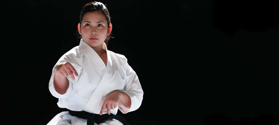 Martial Arts Uniforms 桌布 壁紙 — 一位專注的女性穿著白色空手道服，繫著黑帶，在純黑色背景下擺出強而有力的姿勢。