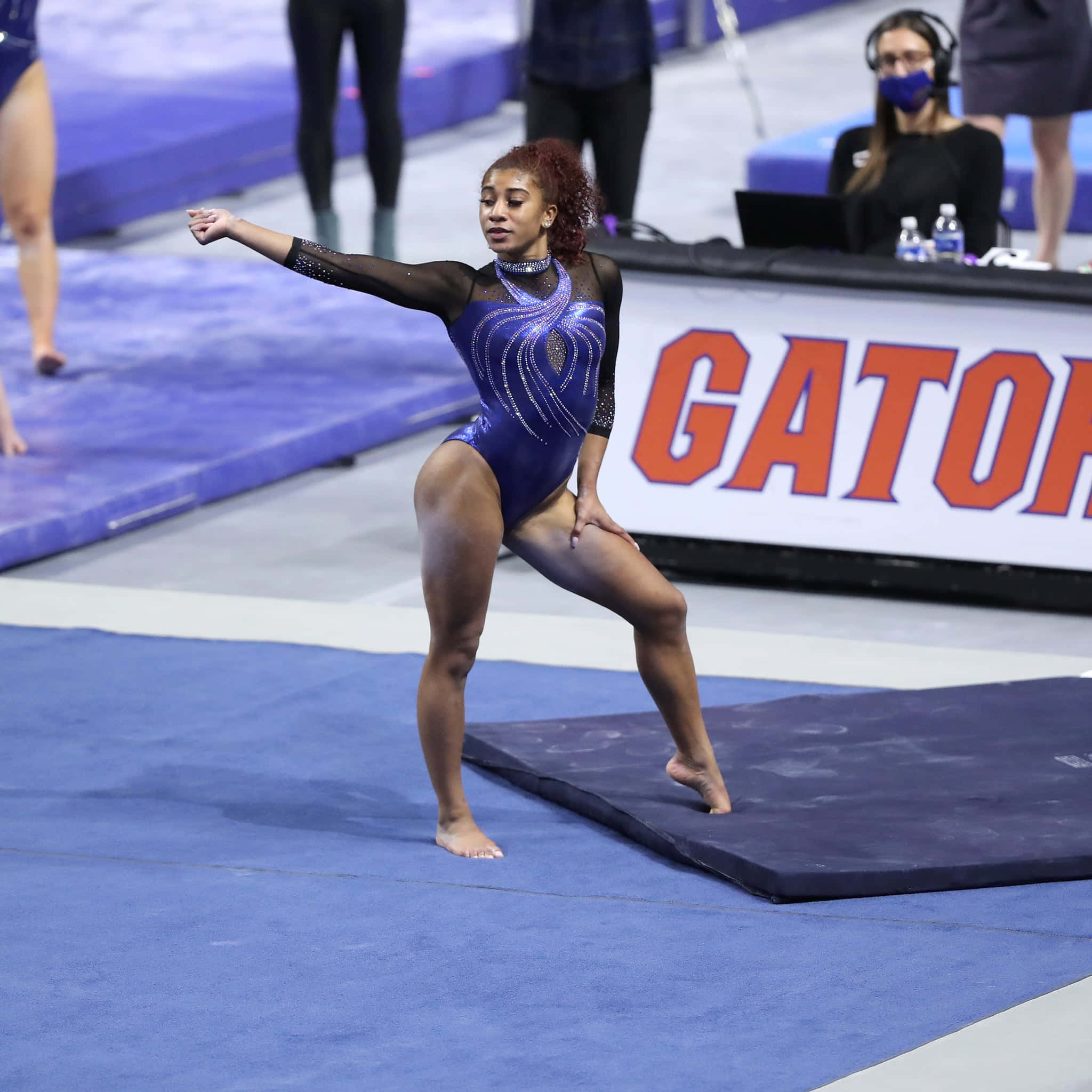 Gymnastics Mat 桌布 壁紙 — 一位專注的女性體操運動員穿著閃亮的藍色緊身衣，在藍色墊子上執行優雅的姿勢，進行競技套路。