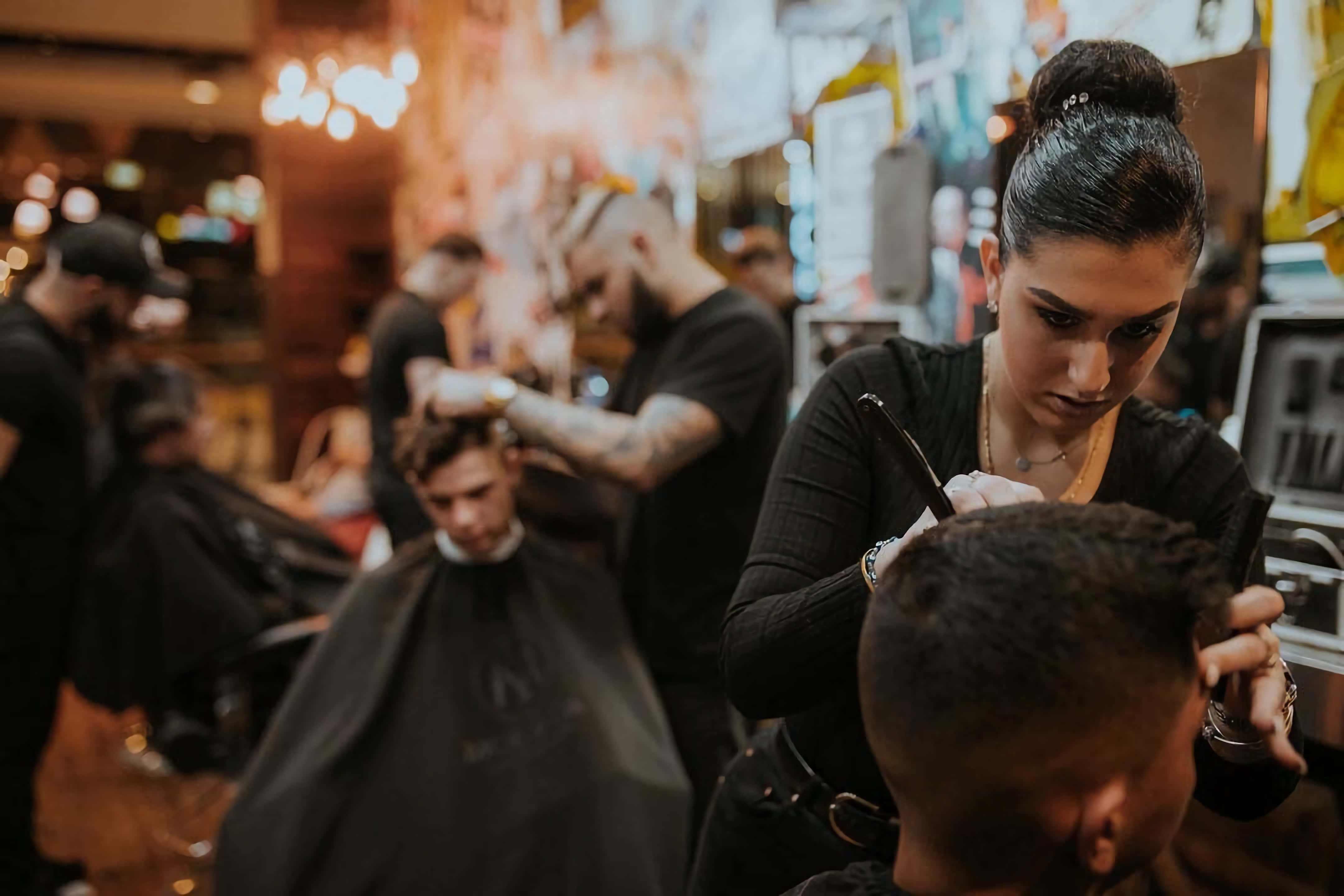 Barber 桌布 壁紙 — 一位專注的女理髮師在燈光溫暖、熙來攘往的理髮店裡，細心地為客人修剪頭髮，店內還有其他造型師和顧客。