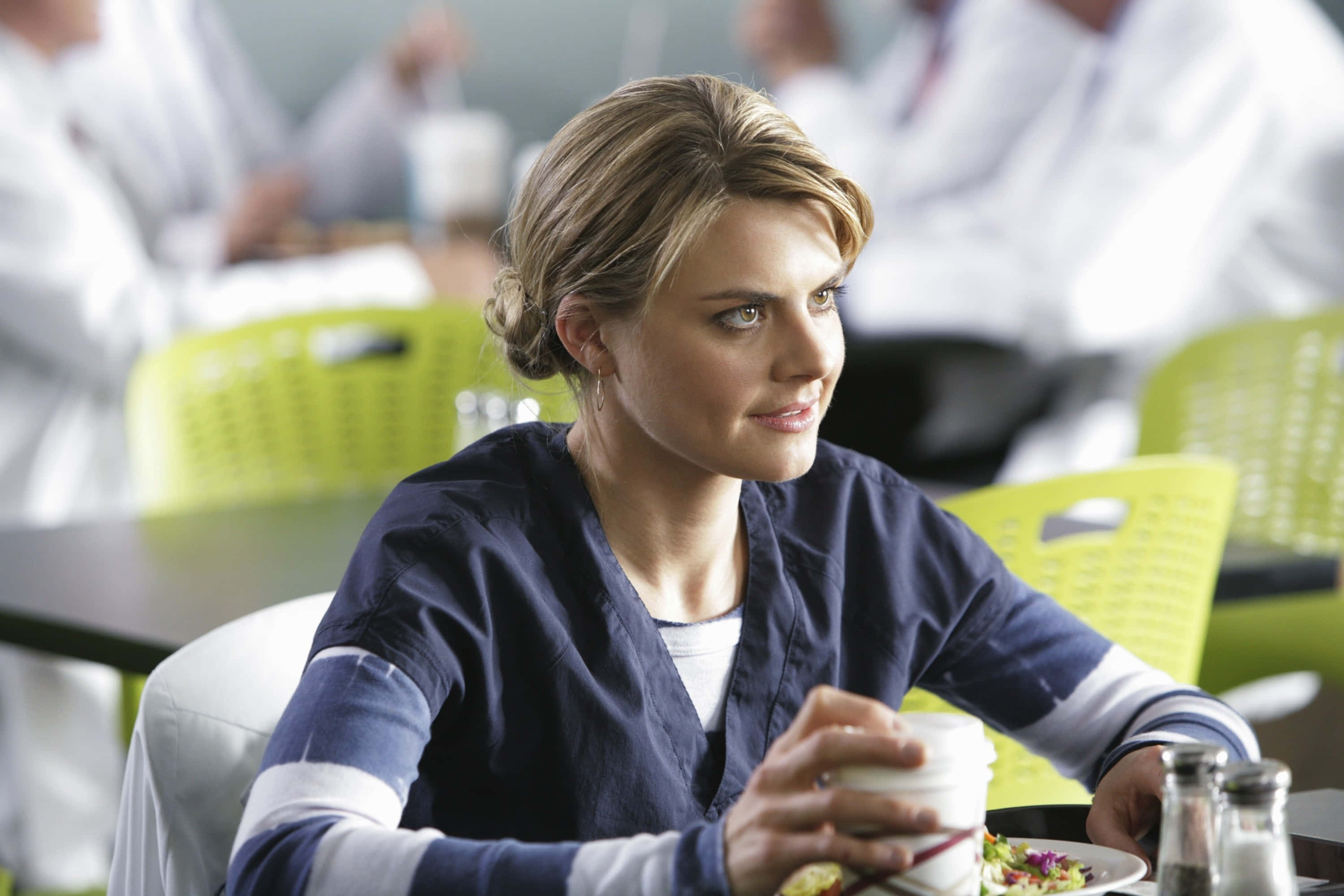 eliza coupe 桌布 壁紙 — 一位專注的女護士穿著藍色手術衣，坐在咖啡廳的桌子旁，手持咖啡杯，身後有模糊的白色實驗室外套同事。