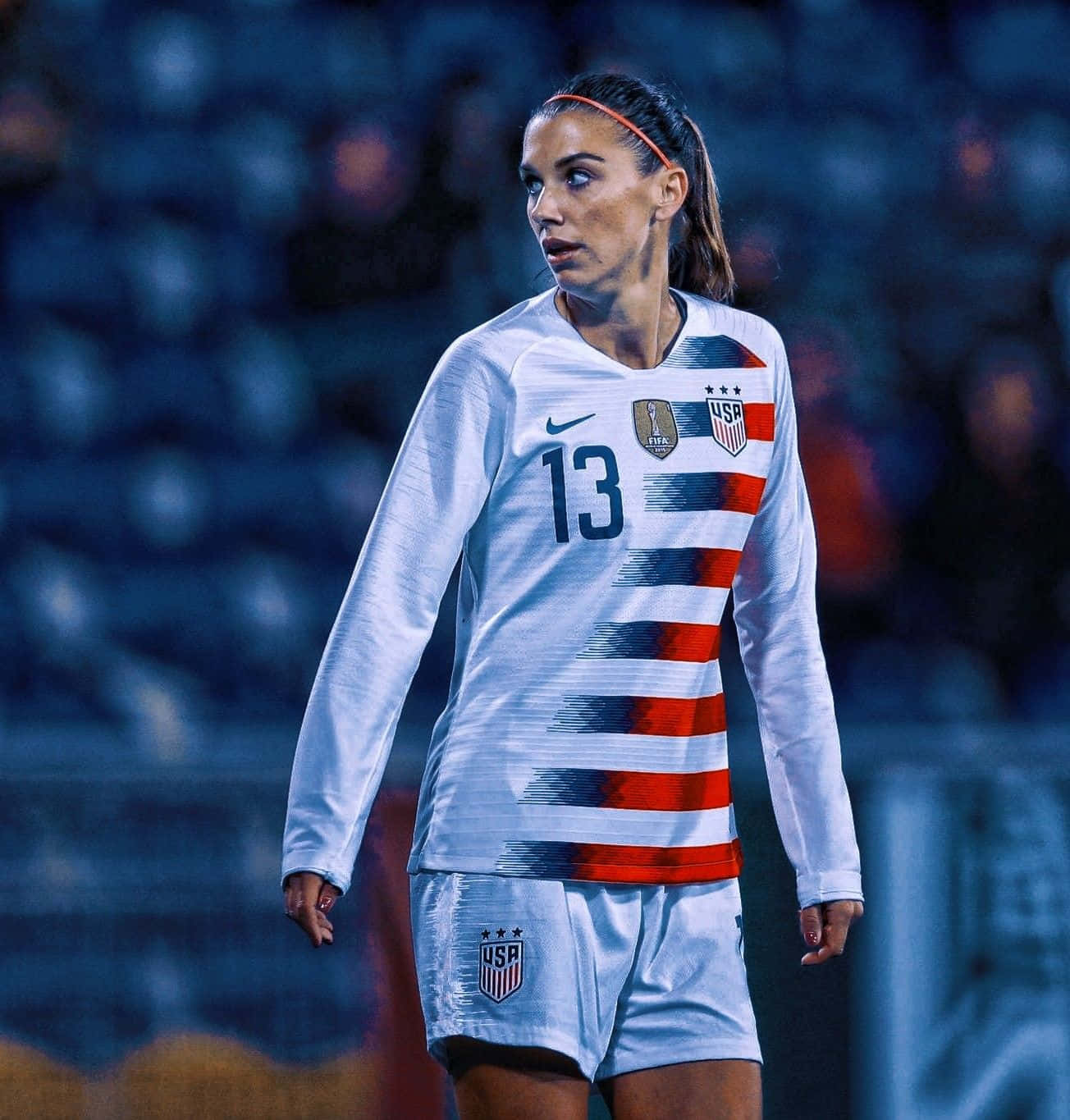 Alex Morgan 桌布 壁紙 — 一位專注的女足球員，身穿帶有紅藍條紋的白色美國國家隊球衣，站在燈光昏暗的球場上。