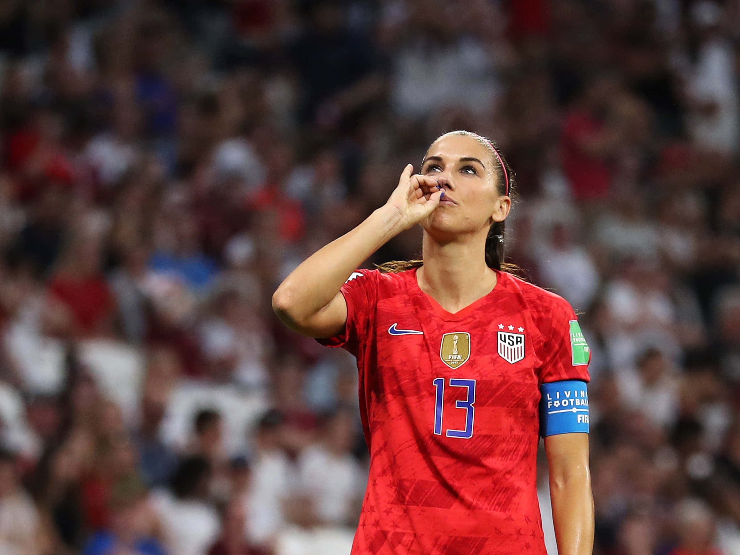Alex Morgan 桌布 壁紙 — 一位專注的女足球員，身穿紅色美國球衣，號碼13，在夜間體育館比賽中若有所思地做手勢。