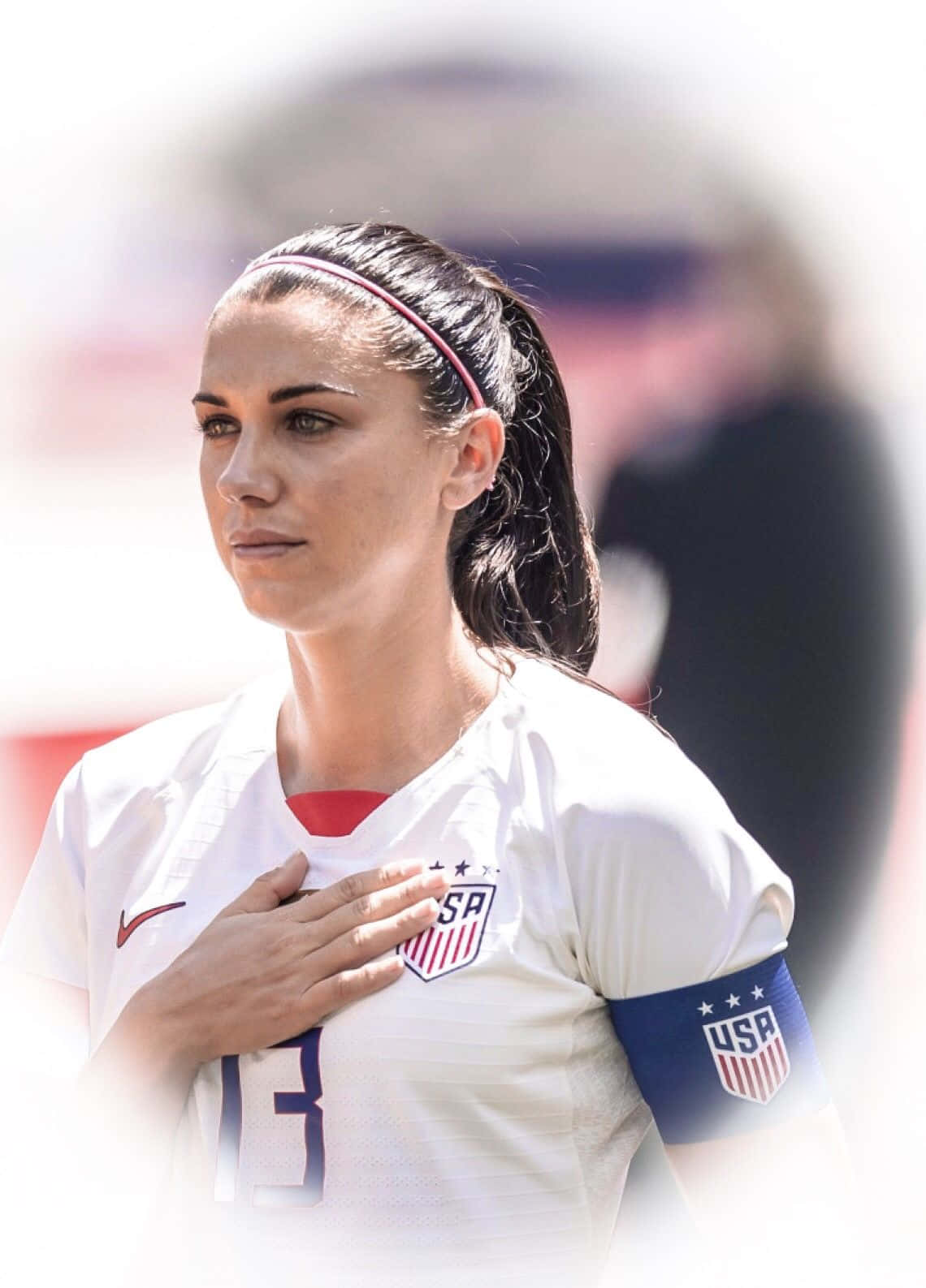 Alex Morgan 桌布 壁紙 — 一位專注的女足球員身穿白色美國球衣，在國歌時刻將手放在心口上。