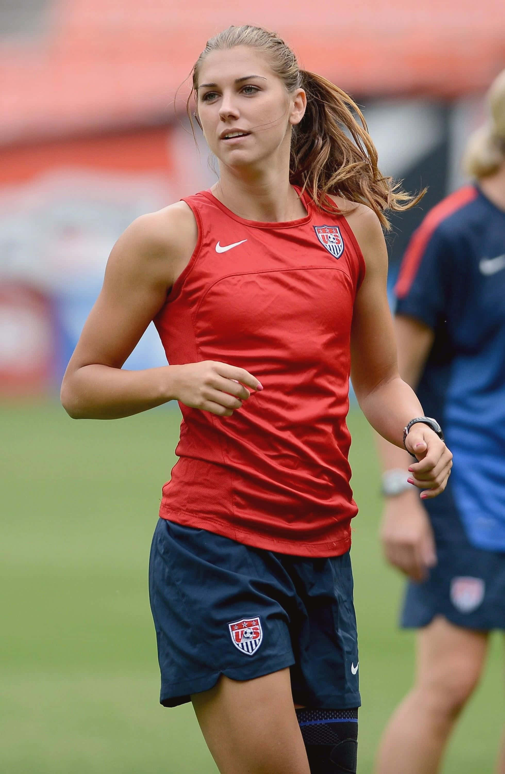 Alex Morgan 桌布 壁紙 — 一位專注的女足球員身穿紅色美國球衣，在日間訓練中衝過綠色球場。