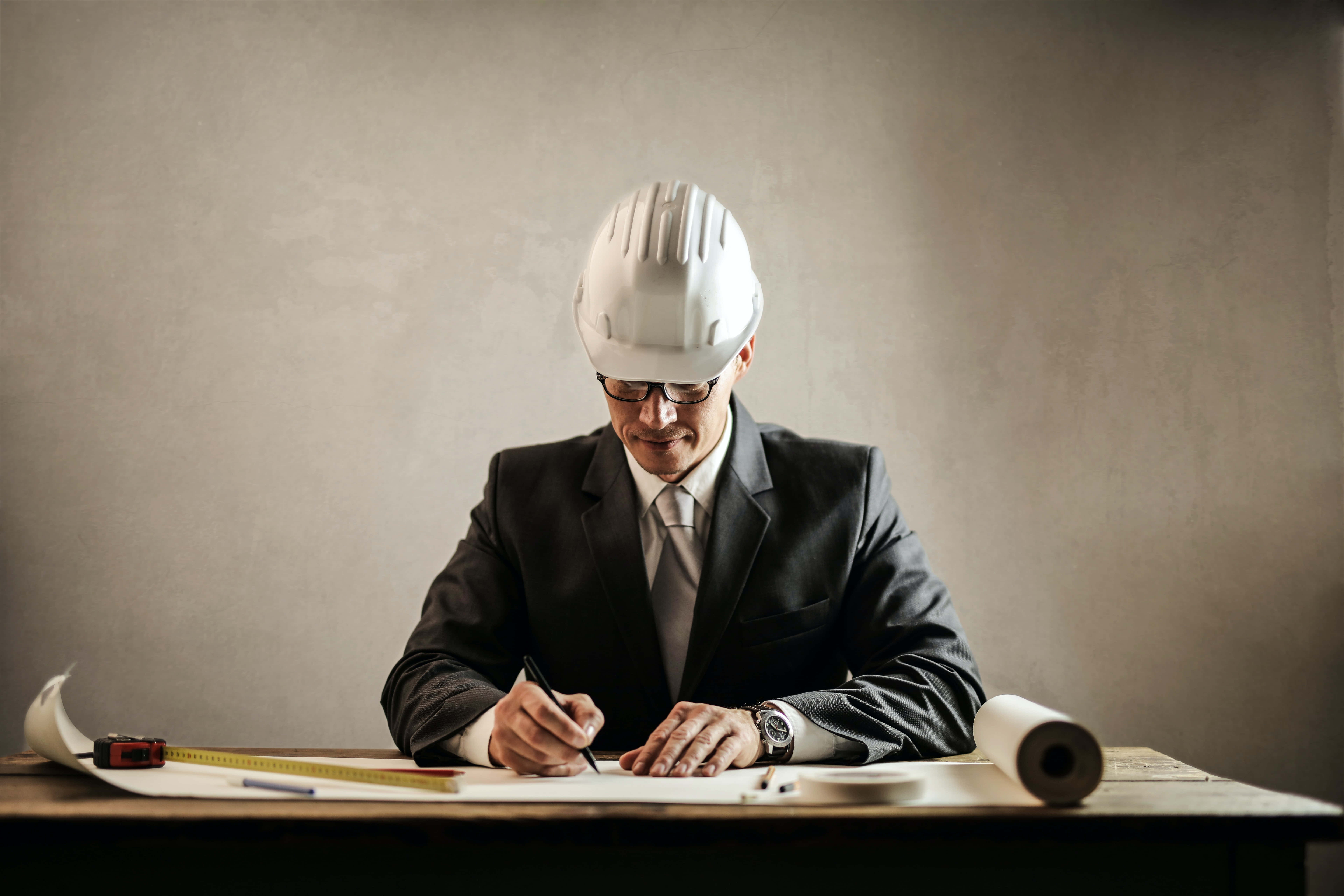 Engineer 桌布 壁紙 — 一位專注的建築師，戴著白色安全帽和西裝，在木桌上簽署藍圖，桌上有工具和捲起的計劃。