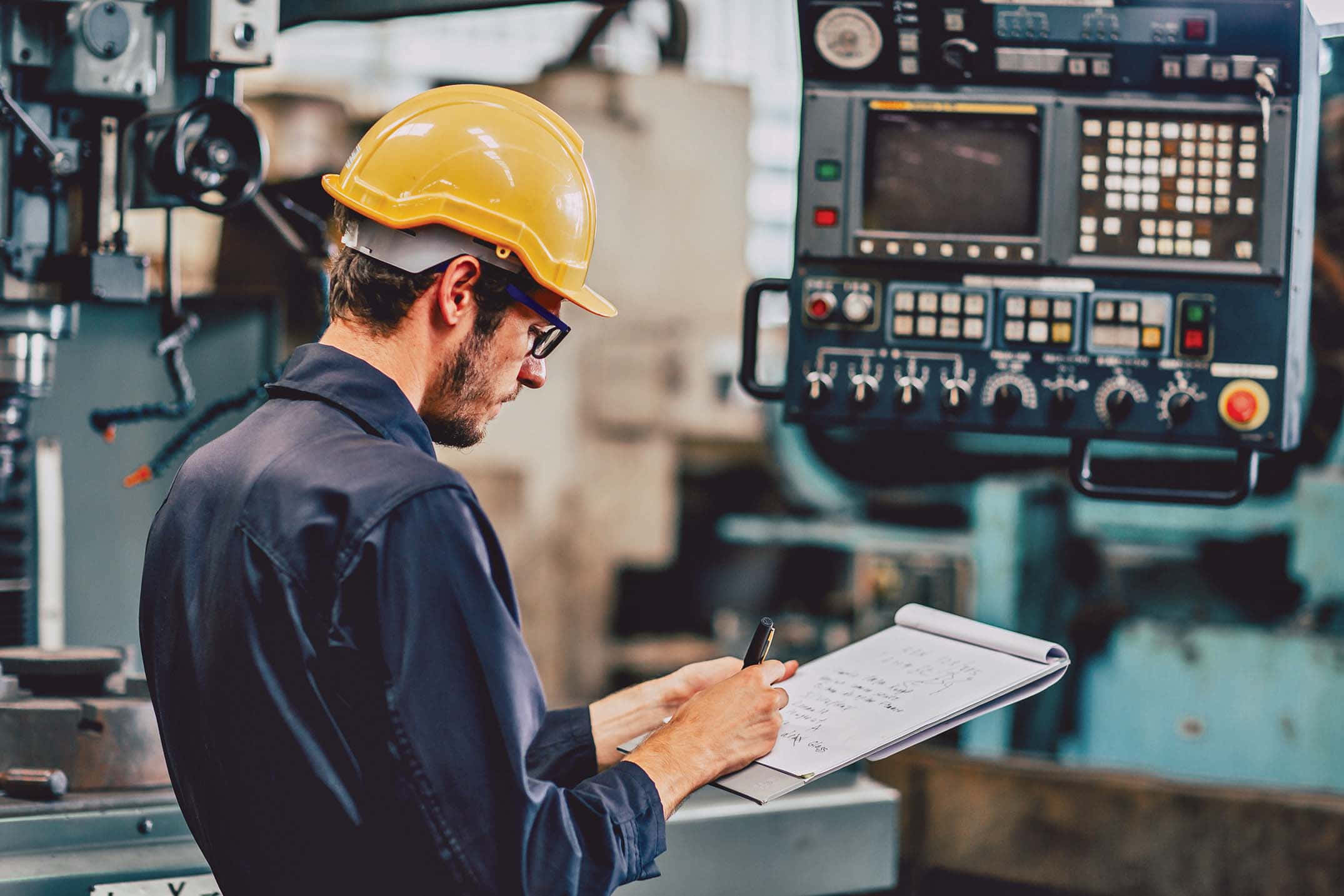 Engineer 桌布 壁紙 — 一位專注的技術人員戴著黃色安全帽，在工廠環境中於複雜的工業機器旁在剪貼板上寫筆記。