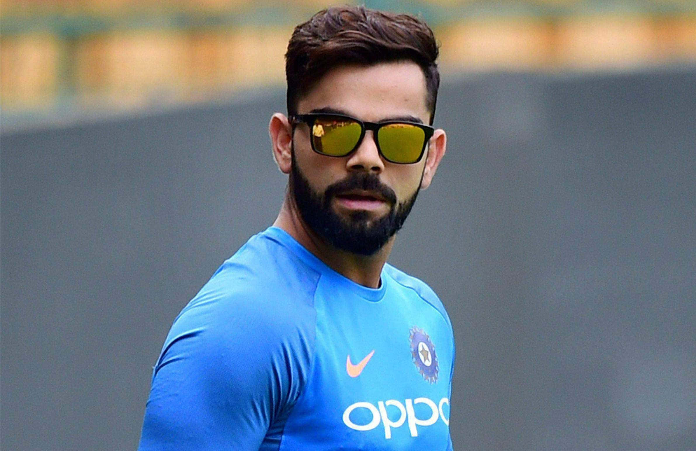 Virat Kohli 桌布 壁紙 — 一位專注的板球運動員穿著藍色訓練球衣和黃色太陽眼鏡，在練習期間專注地凝視。