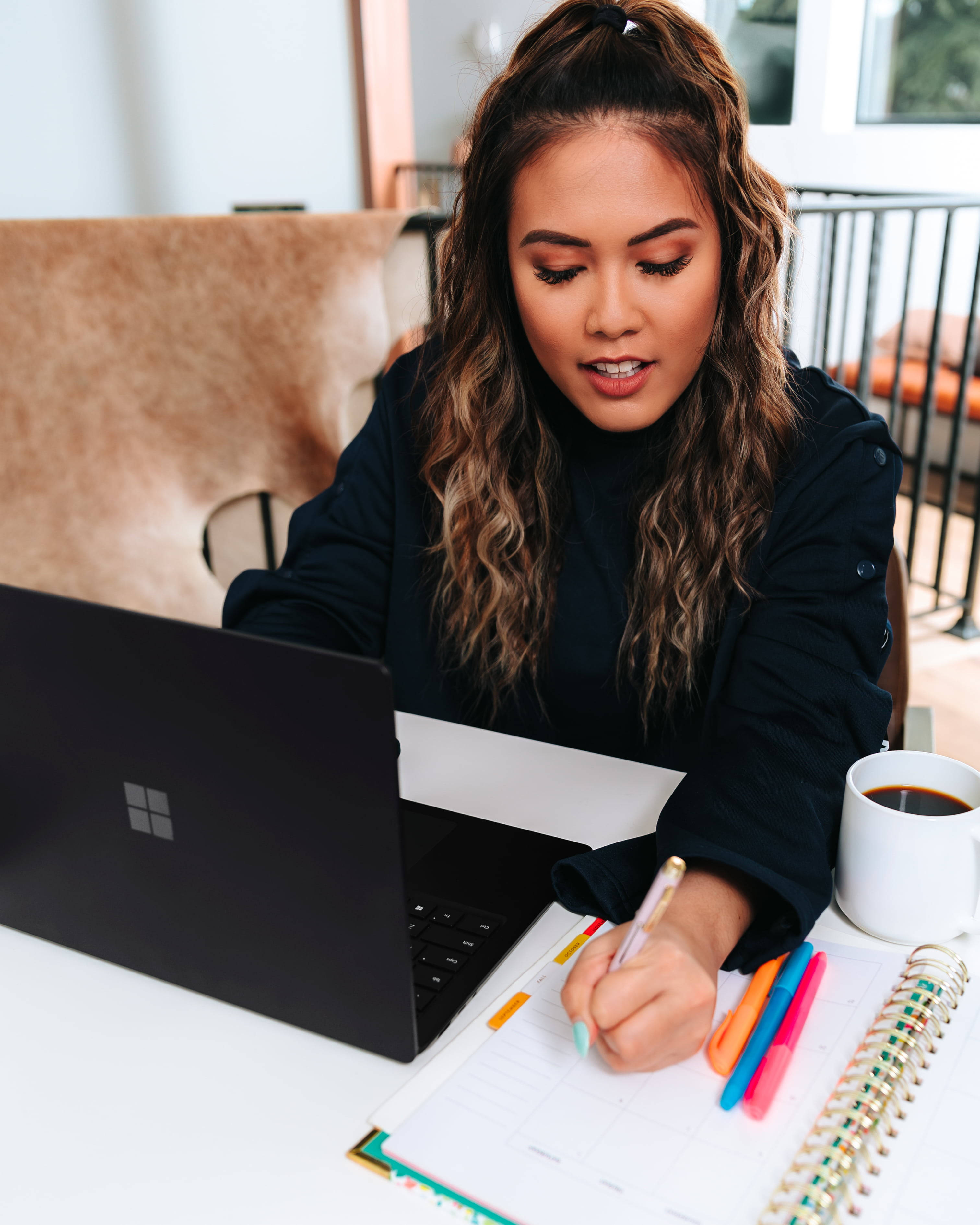 microsoft 桌布 壁紙 — 一位專注的波浪髮女性在明亮的室內環境中，在手帳旁放著筆記型電腦和咖啡杯，正在寫東西。