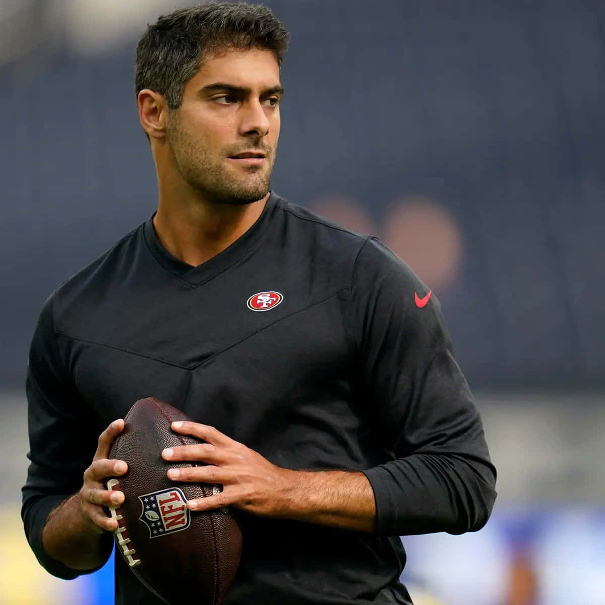 Jimmy Garoppolo 桌布 壁紙 — 一位專注的美式足球員身穿黑色三藩市49人隊球衣，在模糊的球場背景下拿著一個棕色的NFL足球。
