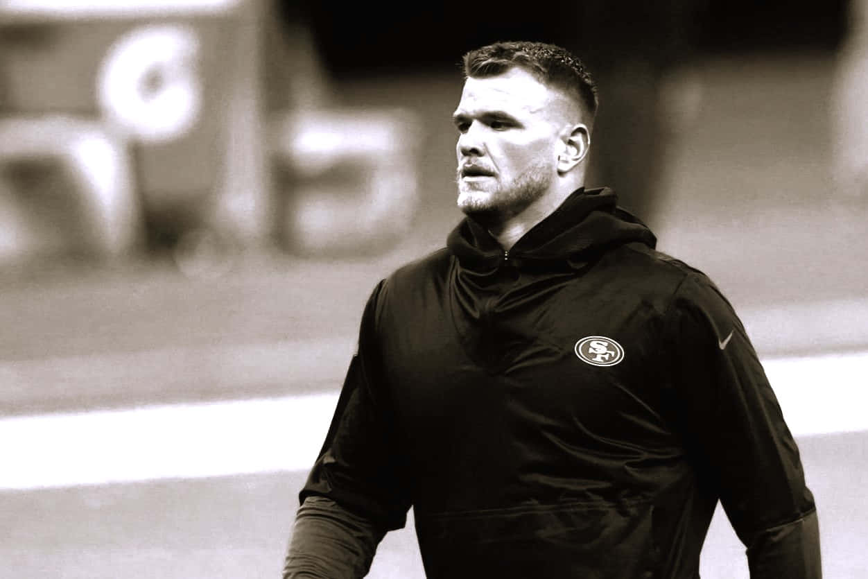 Mike Mcglinchey 桌布 壁紙 — 一位專注的足球員穿著黑色連帽衫，站在球場上，背景是模糊的體育場，呈單色。