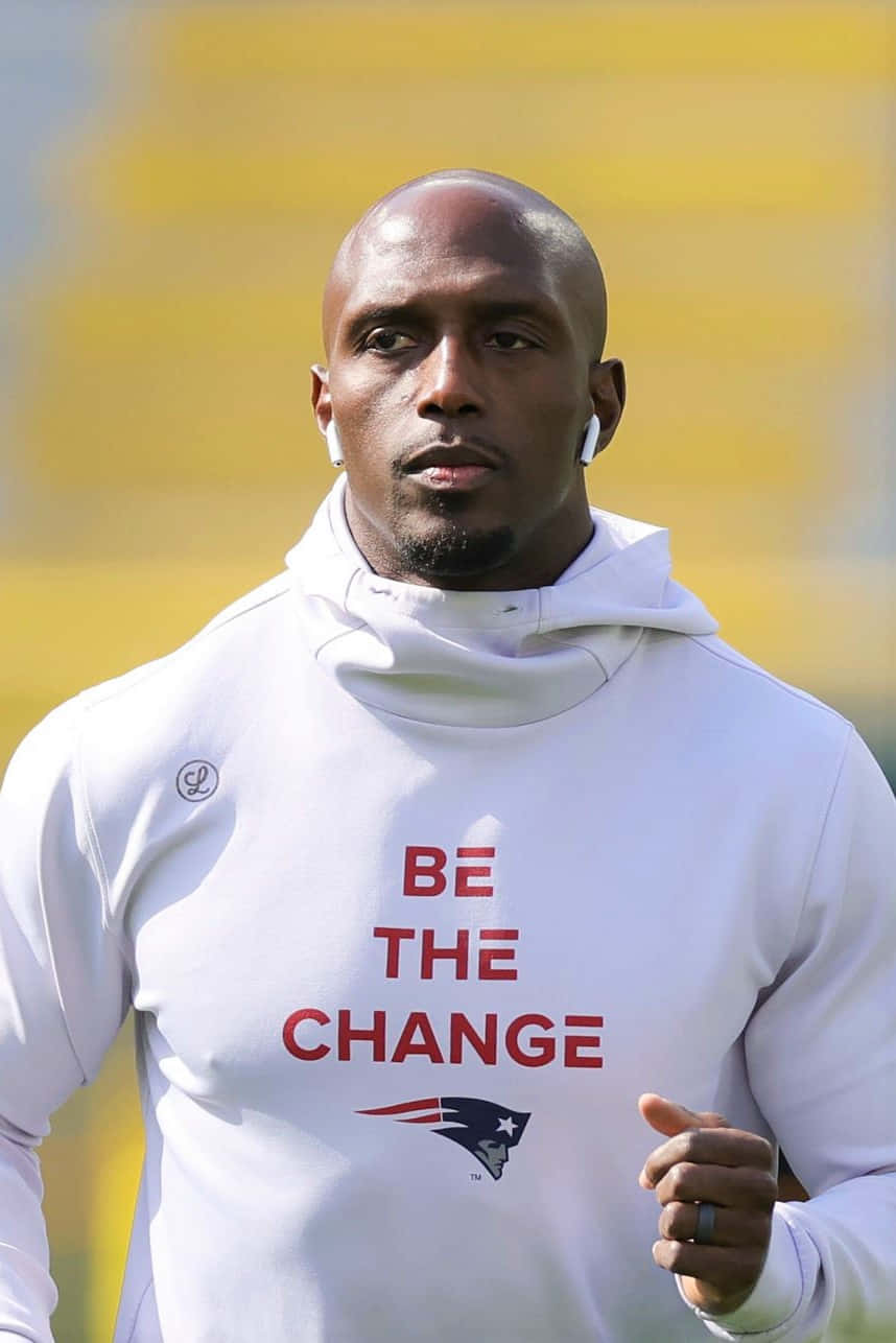 Devin Mccourty 桌布 壁紙 — 一位專注的運動員穿著白色連帽衫，上面有紅色 'BE THE CHANGE' 文字和 Patriots 標誌，站在模糊的黃色體育場背景前。