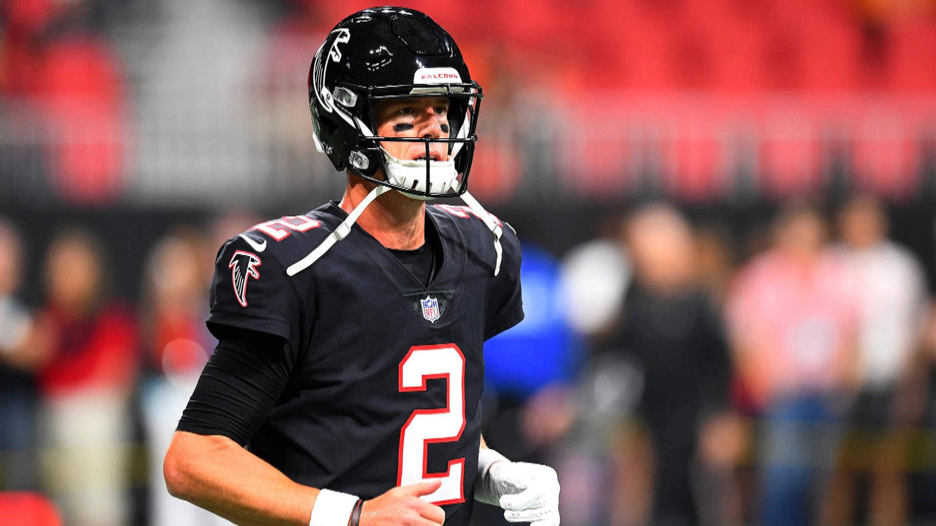 Matt Ryan 桌布 壁紙 — 一位專注的阿特蘭大獵鷹四分衛身穿黑色二號球衣，站在燈火通明的足球場上，身後是模糊的體育場人群。