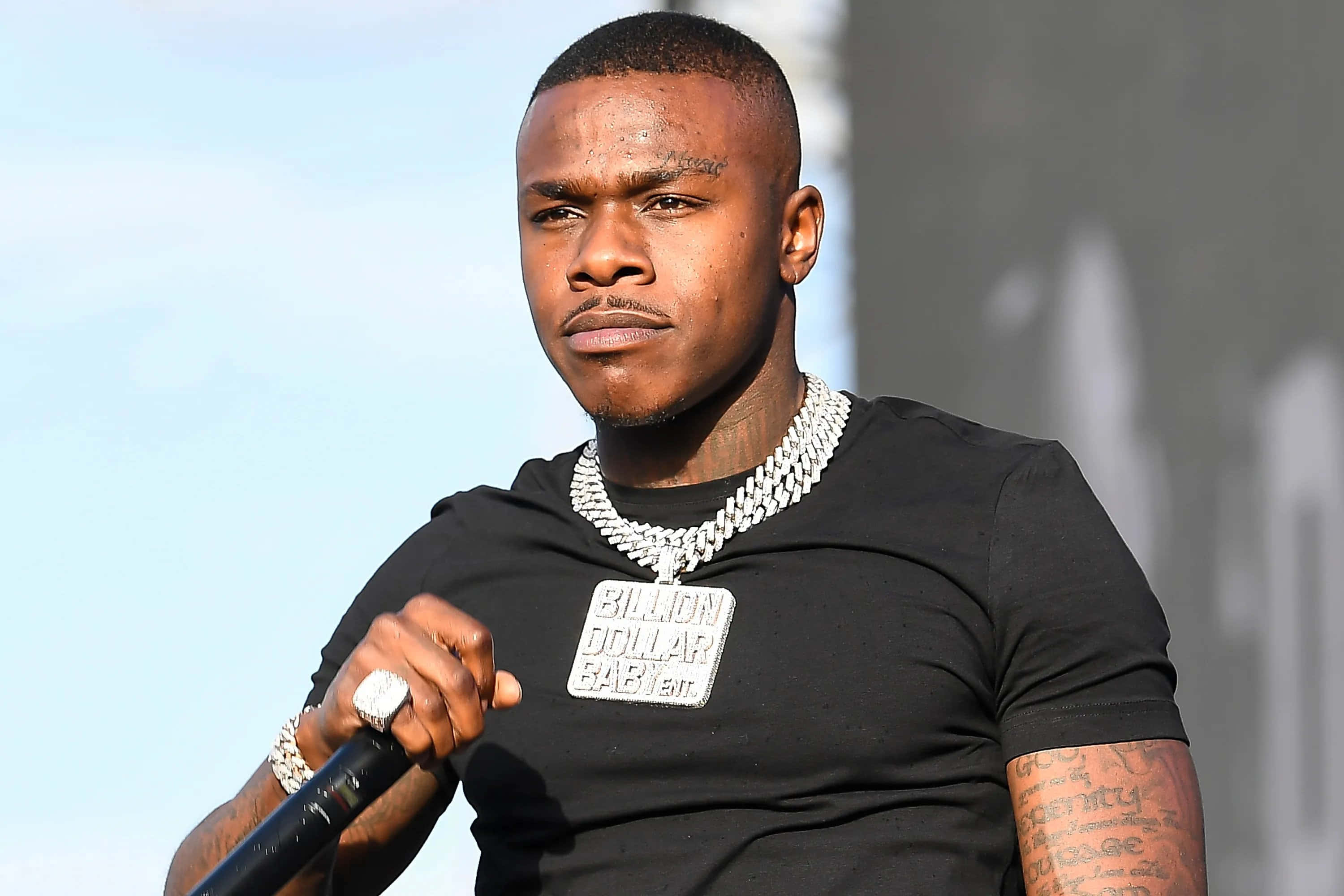 Dababy 桌布 壁紙 — 一位專注的饒舌歌手，身上有紋身和一條粗鏈，在明亮的日光下拿著麥克風在舞台上表演。