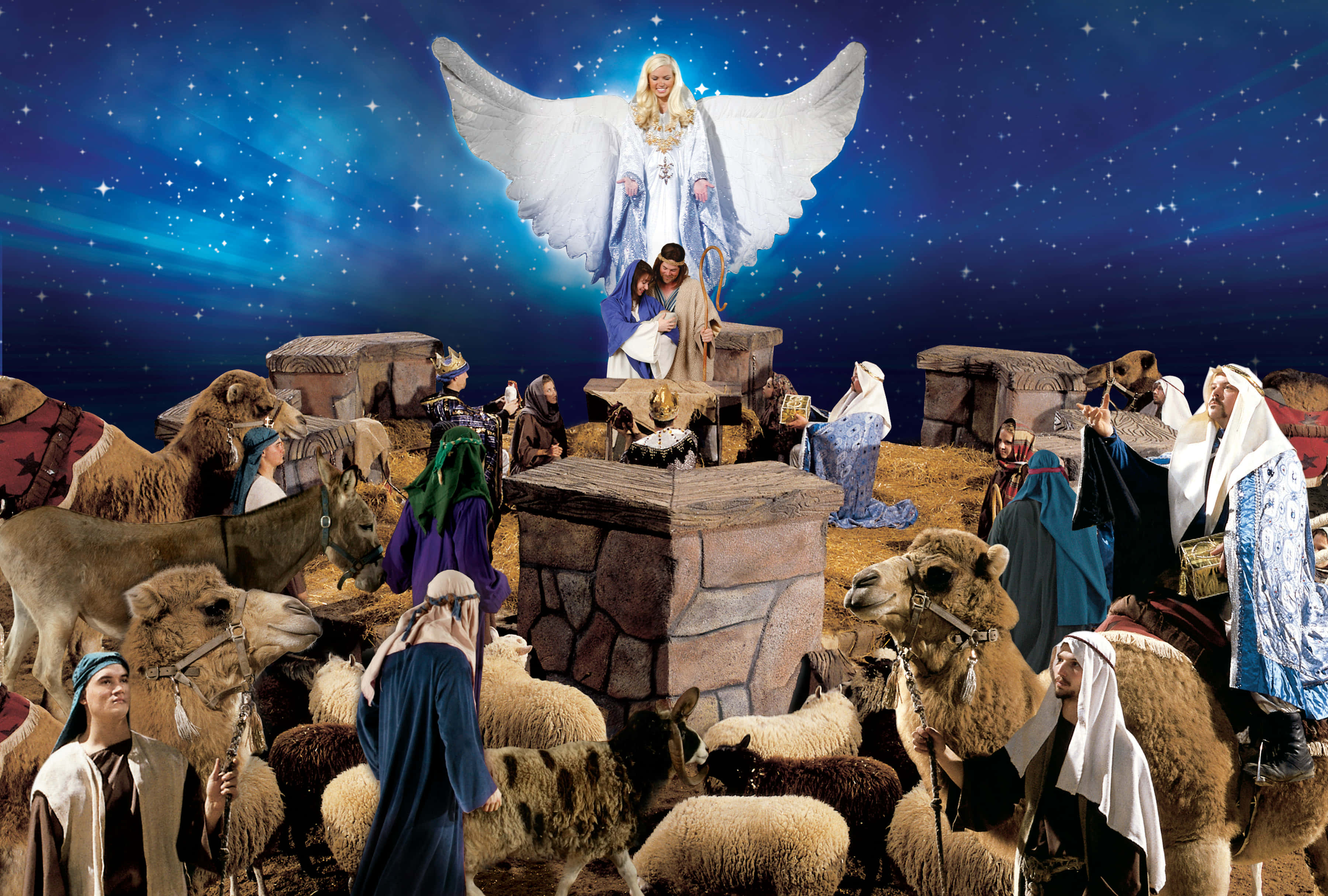 Nativity 桌布 壁紙 — 一位帶有發光翅膀的天使降臨在擠滿駱駝、綿羊和穿著長袍的人物、星空下的耶穌誕生場景之上。