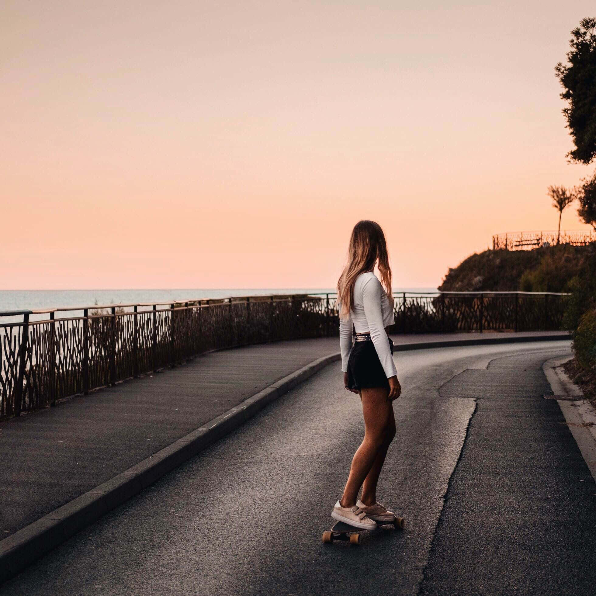 skateboarding 桌布 壁紙 — 一位年輕女子在日落時分沿著海岸公路玩長板，天空呈現溫暖的粉紅色和橙色。
