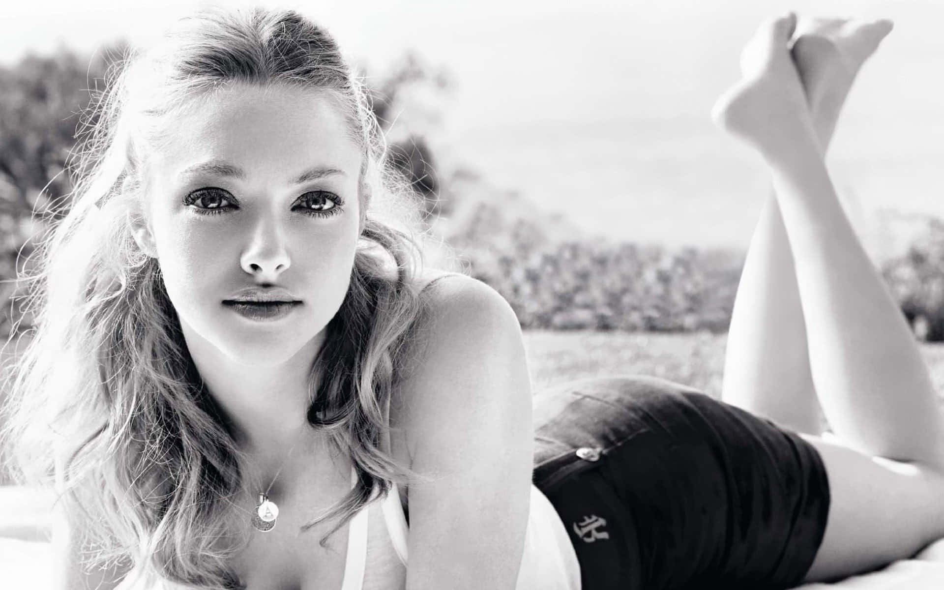 Amanda Seyfried 桌布 壁紙 — 一位年輕女子留著波浪長髮，在戶外黑白相間躺著，雙腿抬起，直視鏡頭。