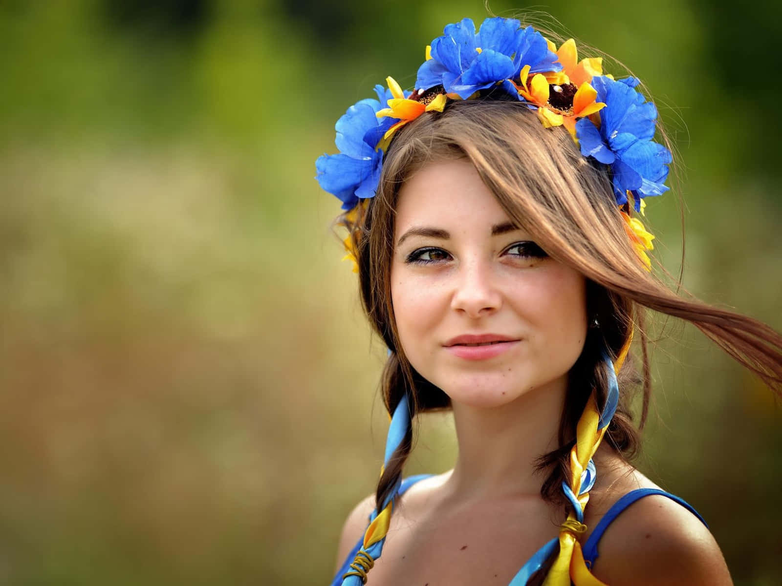Ukrainian Girl Blue Flowery Headdress 壁紙 — 一位年輕女子編著辮子，戴著鮮豔的藍黃色花冠，背景是柔和的模糊綠色。
