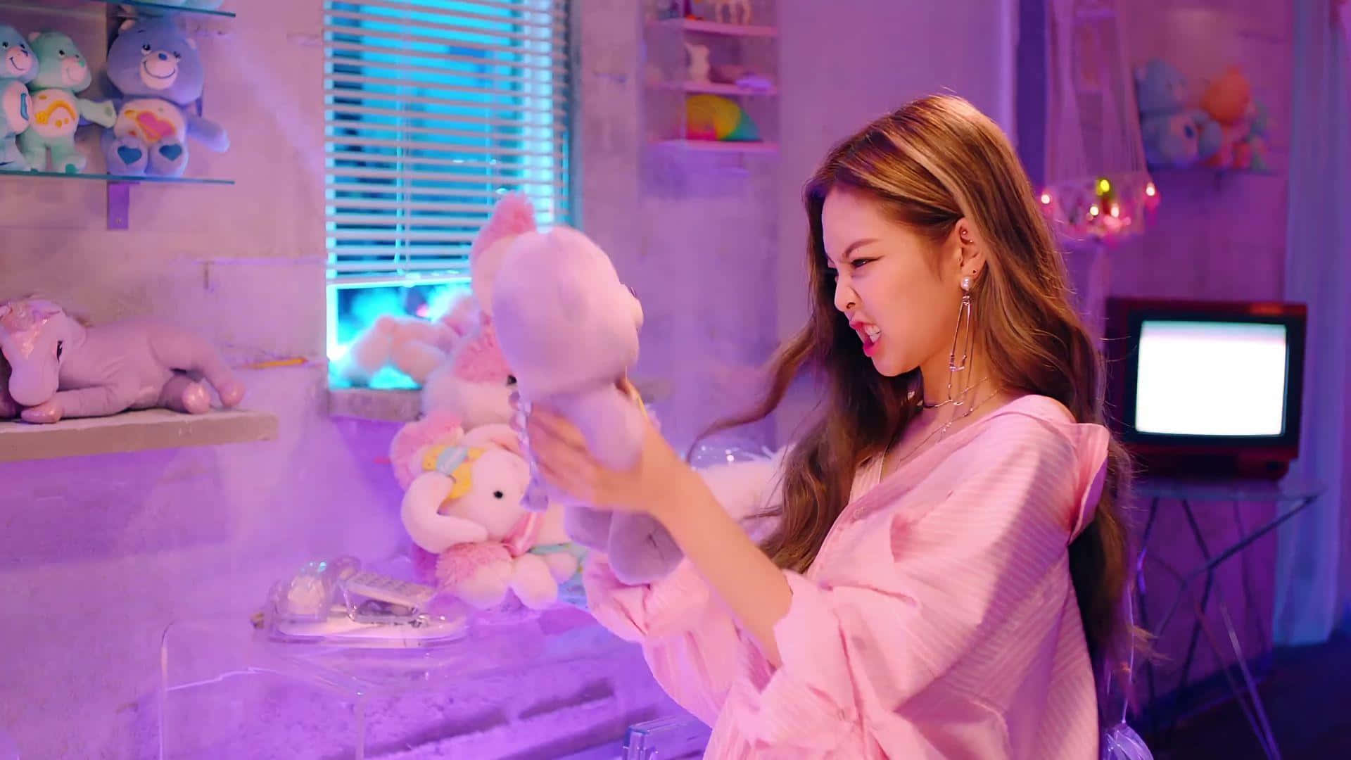 Jennie Kim 桌布 壁紙 — 一位年輕女子身穿粉紅色睡袍，在充滿毛絨玩具的紫色燈光房間裡，俏皮地抱著一個毛絨獨角獸。