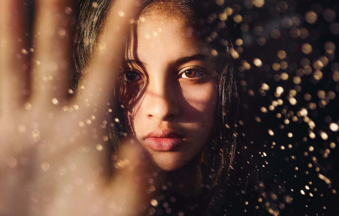 Brown Eyed Girl 桌布 壁紙 — 一位年輕女子透過濺滿雨水的窗戶凝視，背景中金色的散景燈光柔和地閃爍。