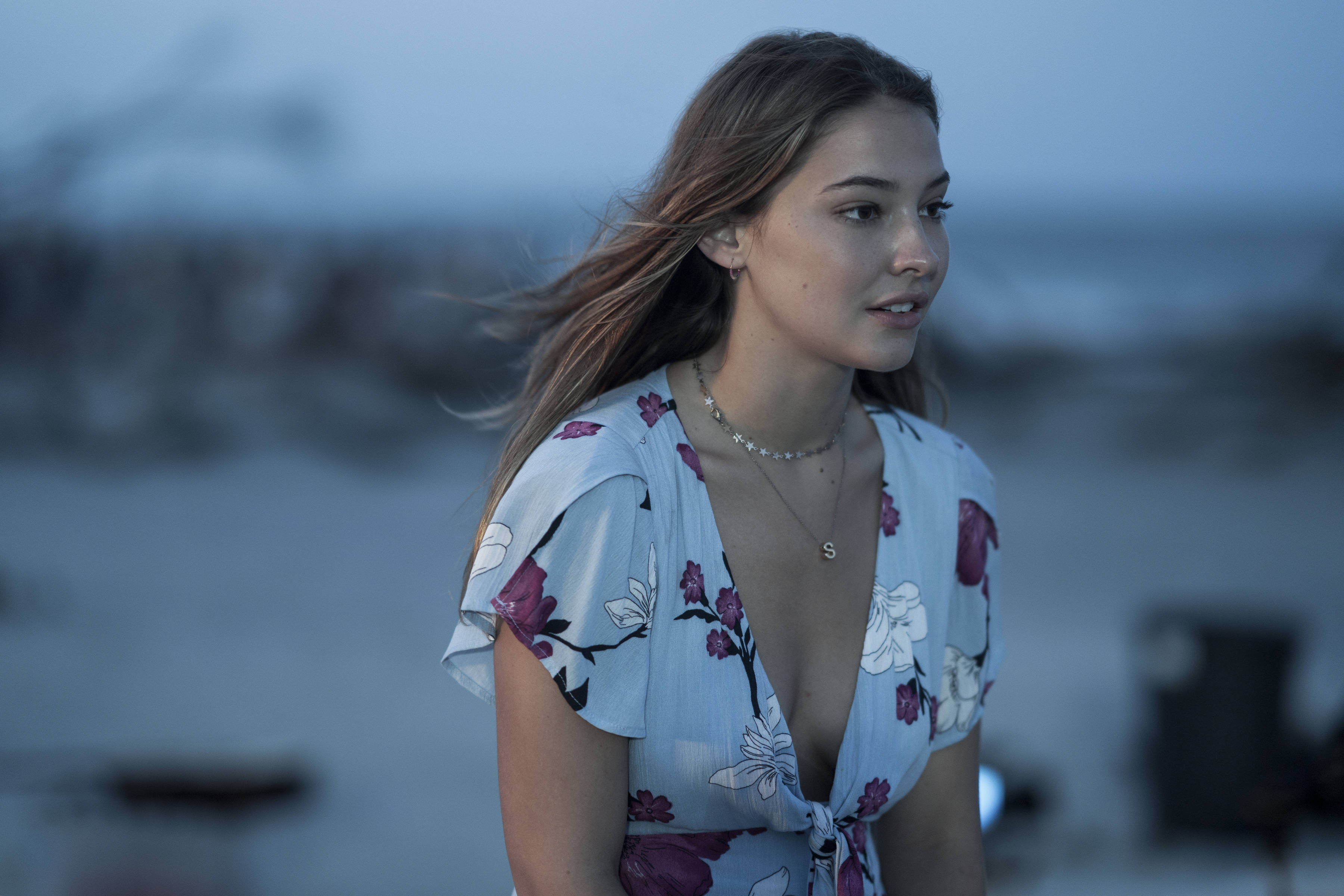 《Outer Banks》Madelyn Cline 壁紙 — 一位年輕女子頭髮被風吹起，在涼爽的暮色天空下若有所思地凝視著地平線。