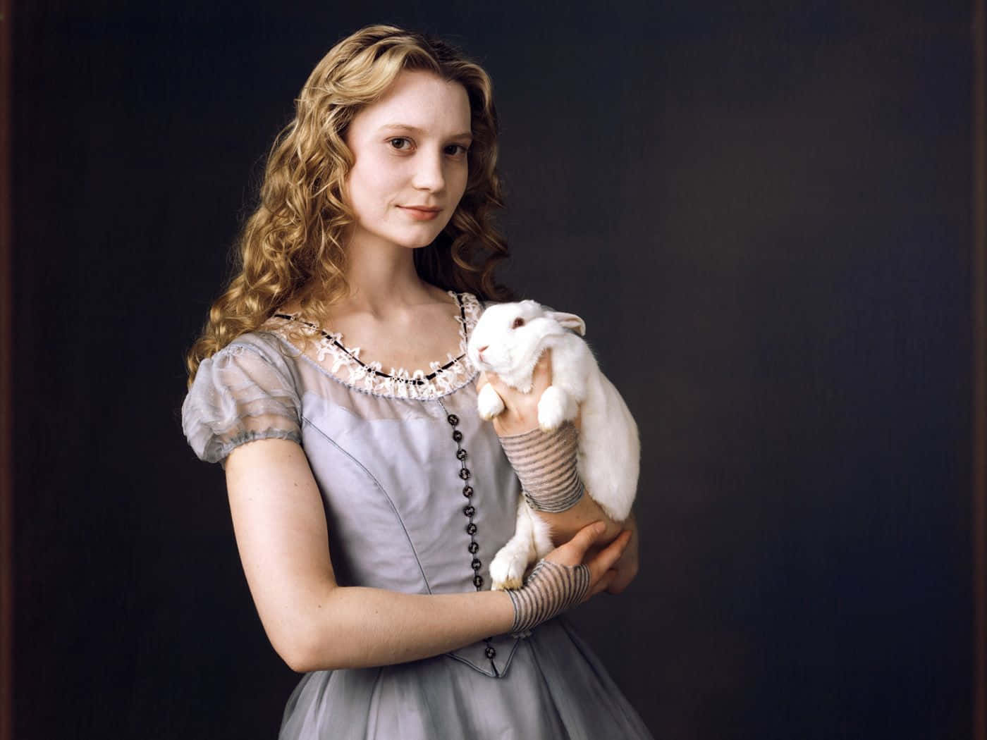 Mia Wasikowska 桌布 壁紙 — 一位年輕女性，一頭捲曲金髮，身穿復古灰色連衣裙，在深色背景前抱著一隻白兔。