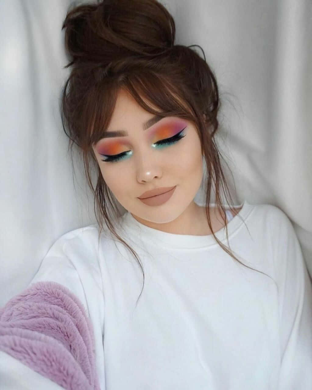 Spring Makeup 桌布 壁紙 — 一位年輕女性，帶有充滿活力的彩虹眼影和凌亂的髮髻，靠在白色布料上，穿著白色襯衫和紫色毛絨袖子。