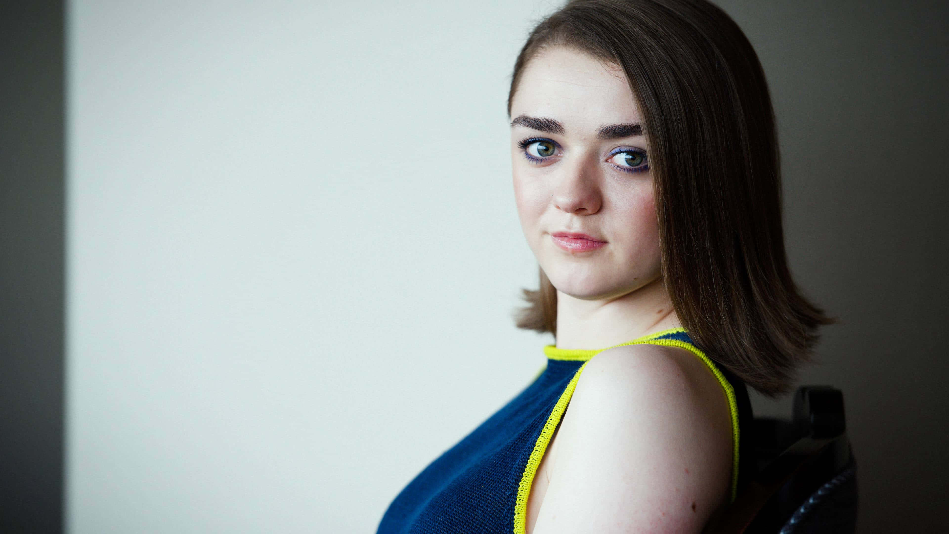 maisie williams 桌布 壁紙 — 一位年輕女性，擁有引人注目的藍色眼睛和深色頭髮，身穿帶有黃色飾邊的藍色上衣，坐在椅子上，背景為純色。
