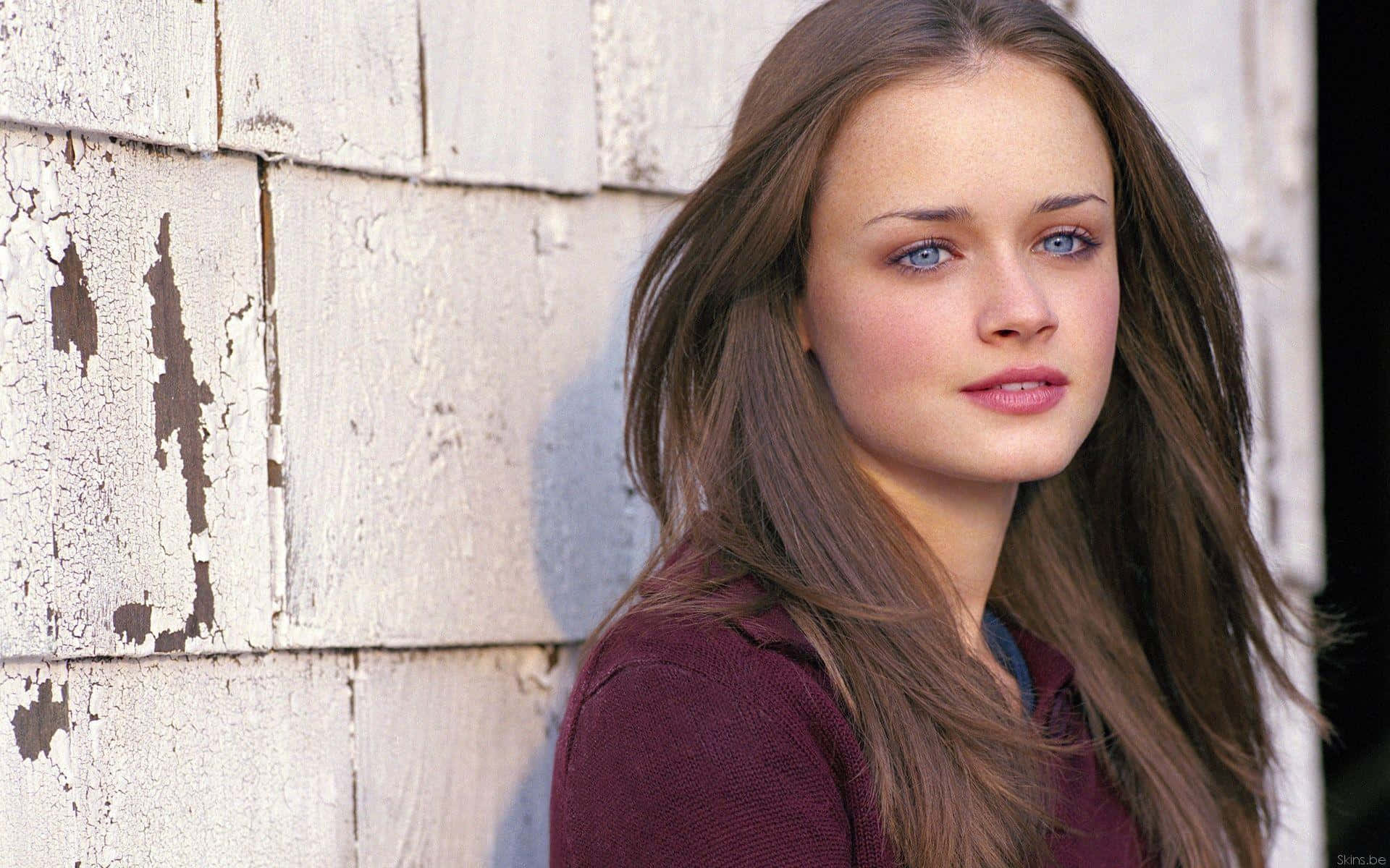 Alexis Bledel 桌布 壁紙 — 一位年輕女性，擁有引人注目的藍色眼睛和長長的棕色頭髮，靠在一面風化的白色木牆上。