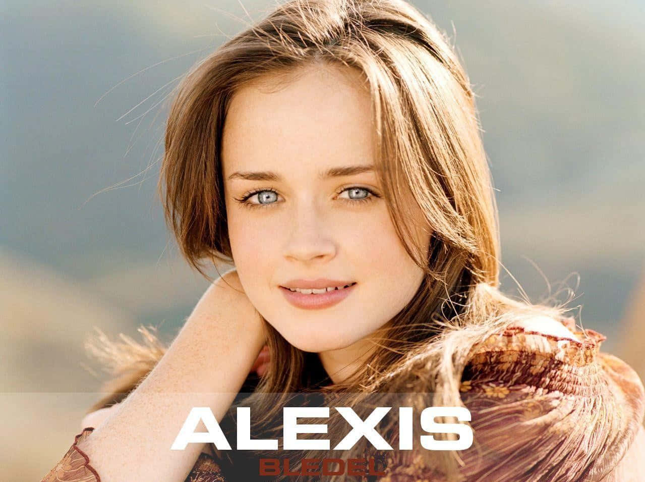 Alexis Bledel 桌布 壁紙 — 一位年輕女性，擁有引人注目的藍色眼睛和陽光親吻的棕色頭髮，在模糊的自然背景下溫柔微笑。