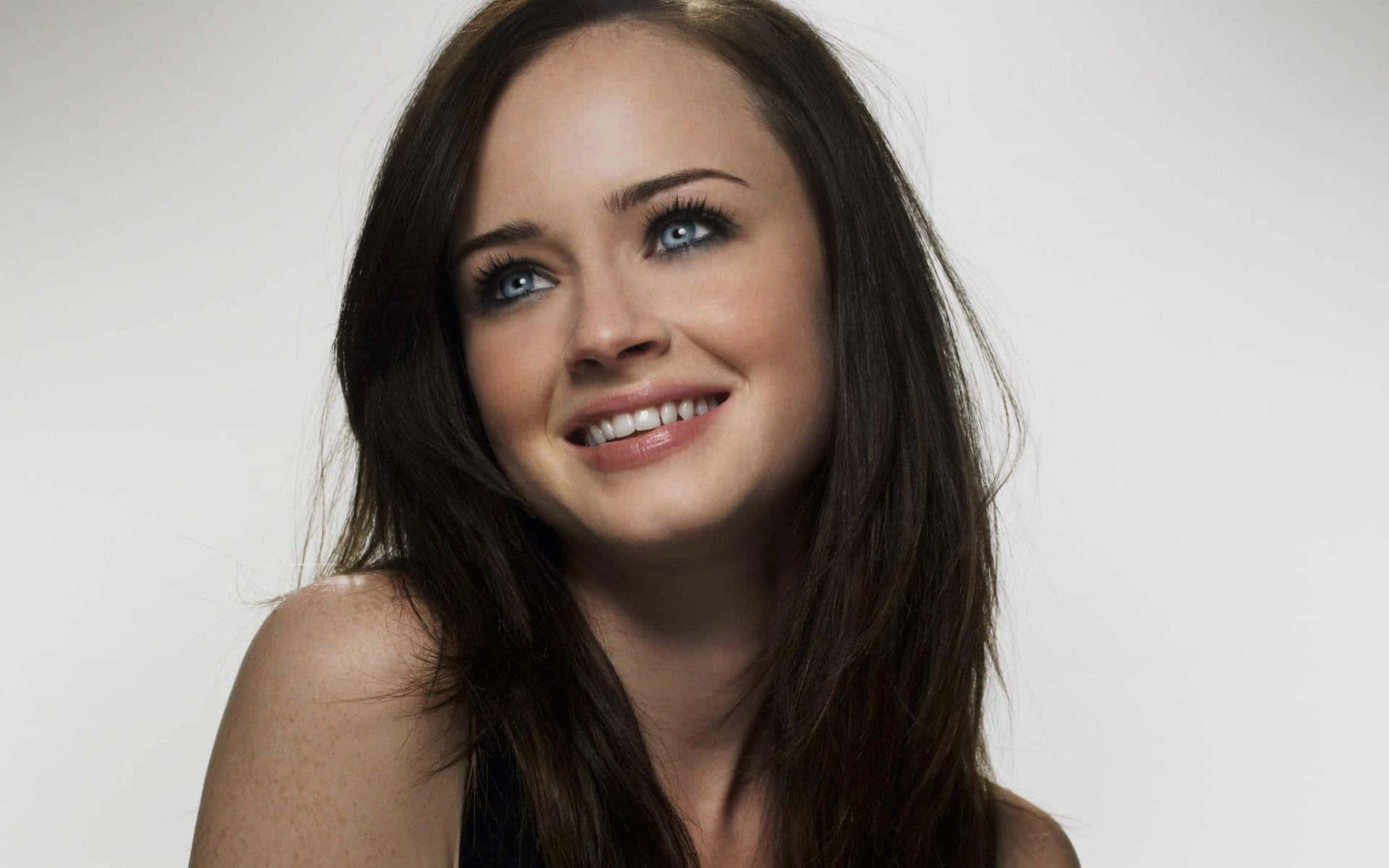 Alexis Bledel 桌布 壁紙 — 一位年輕女性，擁有引人注目的藍色眼睛和深色頭髮，在純白背景前溫暖地微笑。